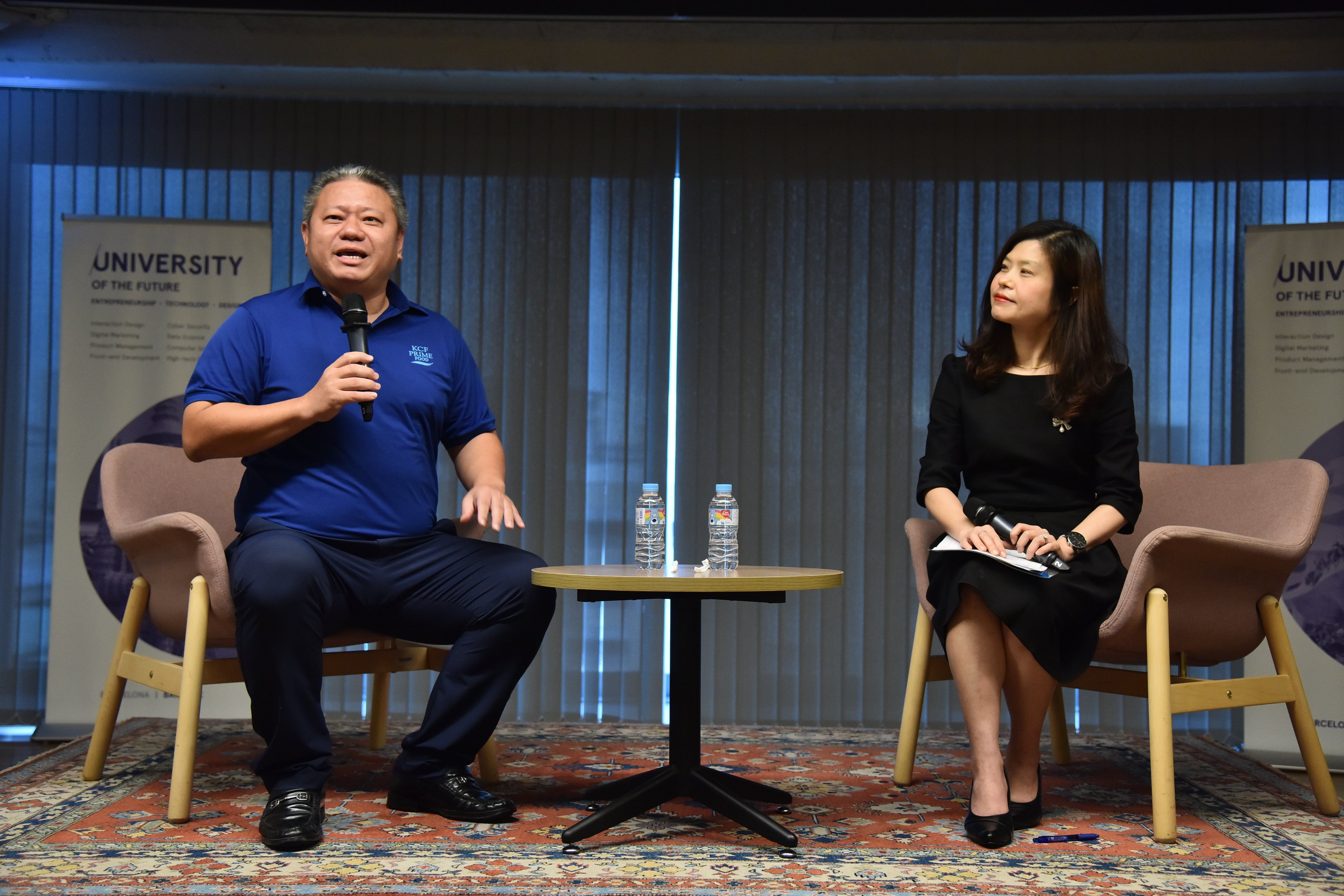A man speaks into a microphone while a woman listens during a presentation.