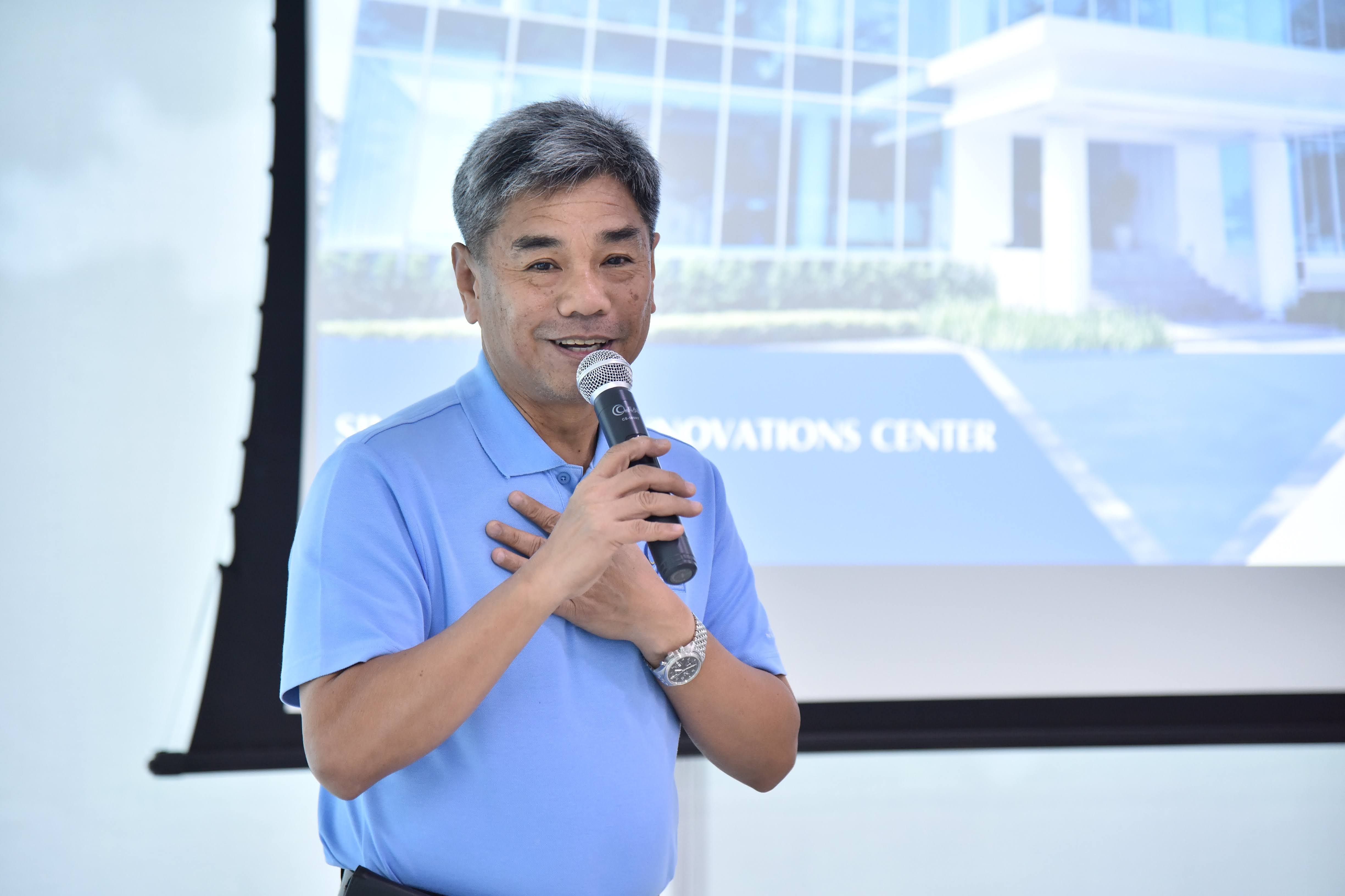 Smiling man in blue shirt speaks into microphone, hand on chest, with projected image.