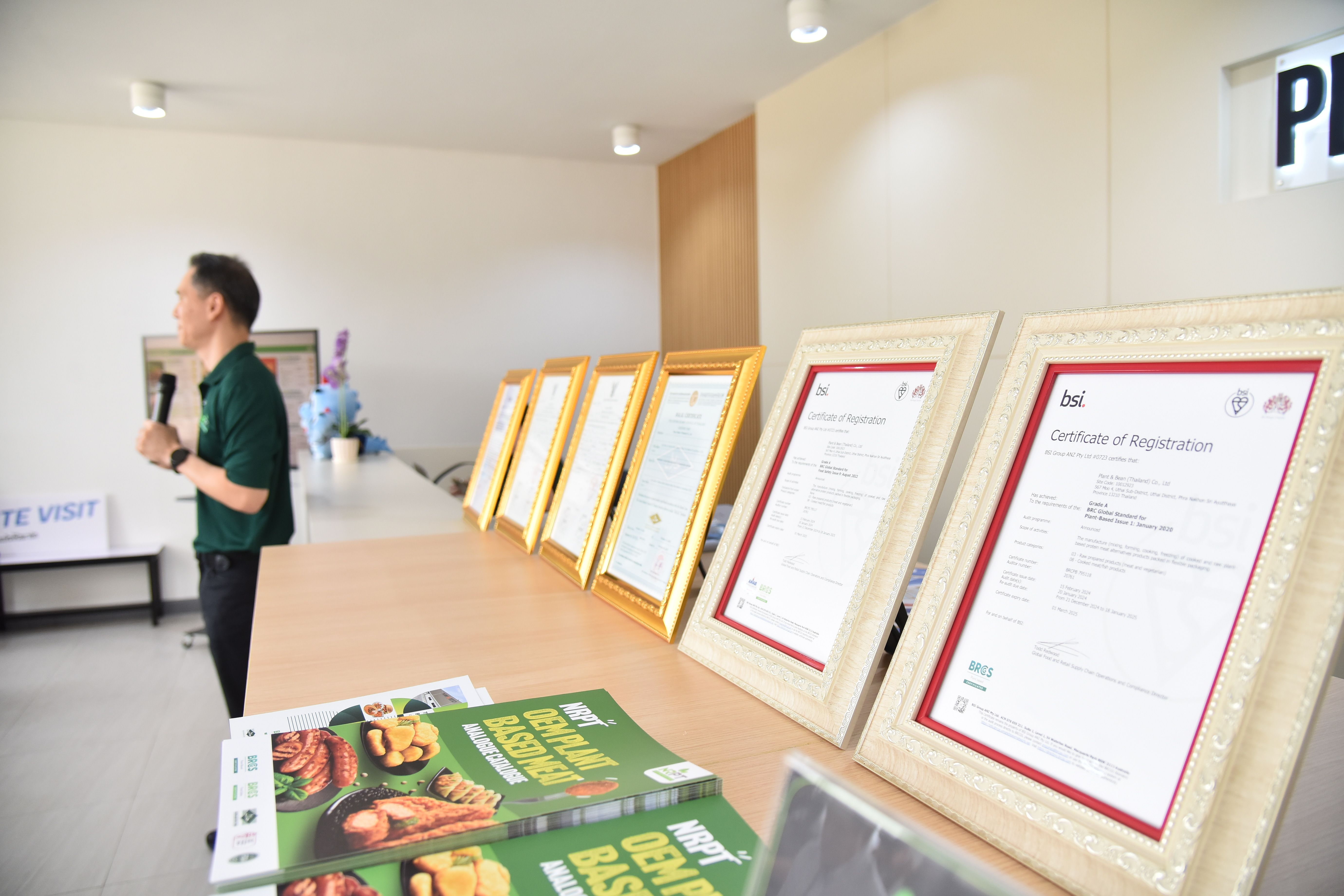 A person speaks at an event while framed certificates are displayed on a table.