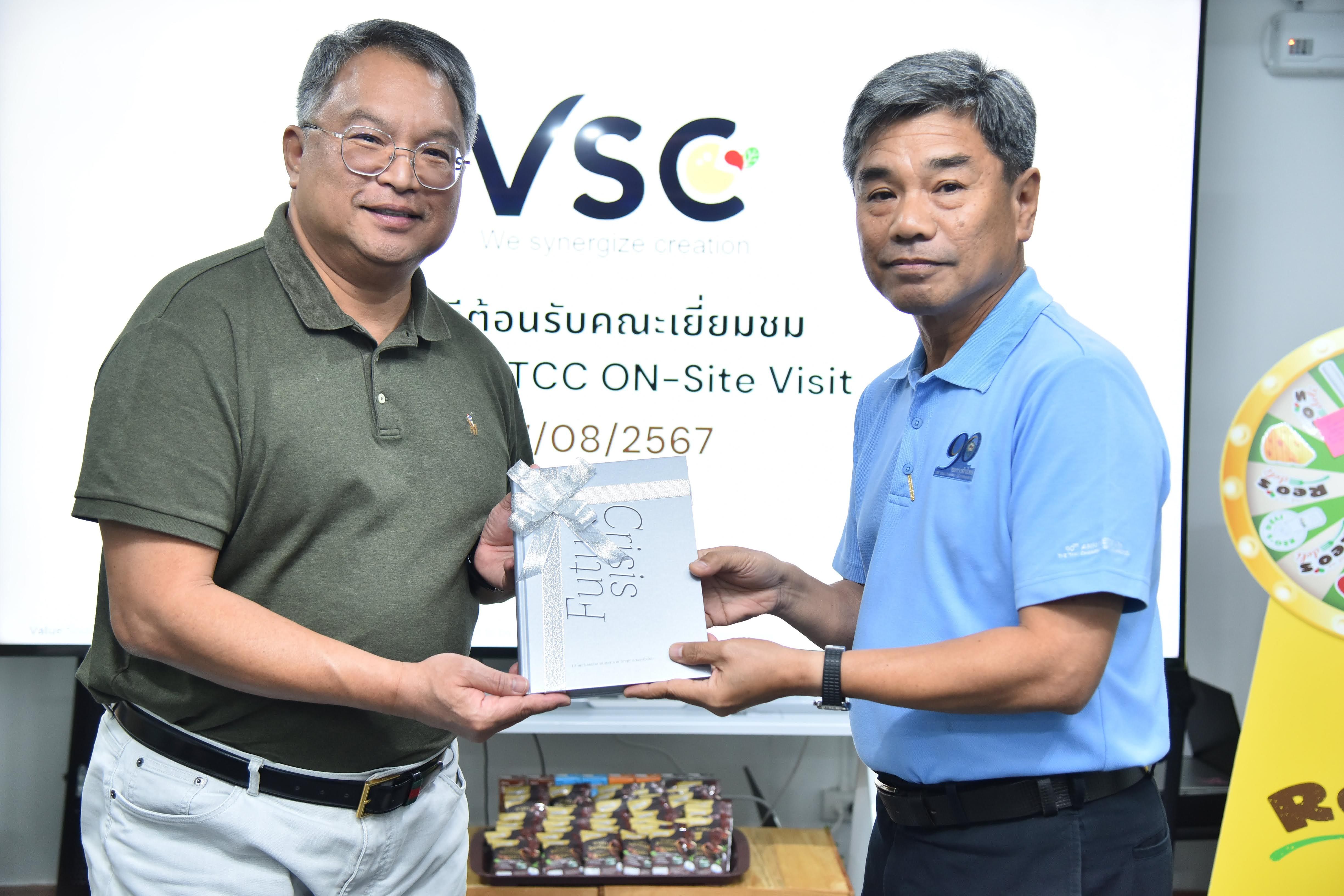 Two men exchange a document in front of a screen displaying 'TCC ON-Site Visit'.