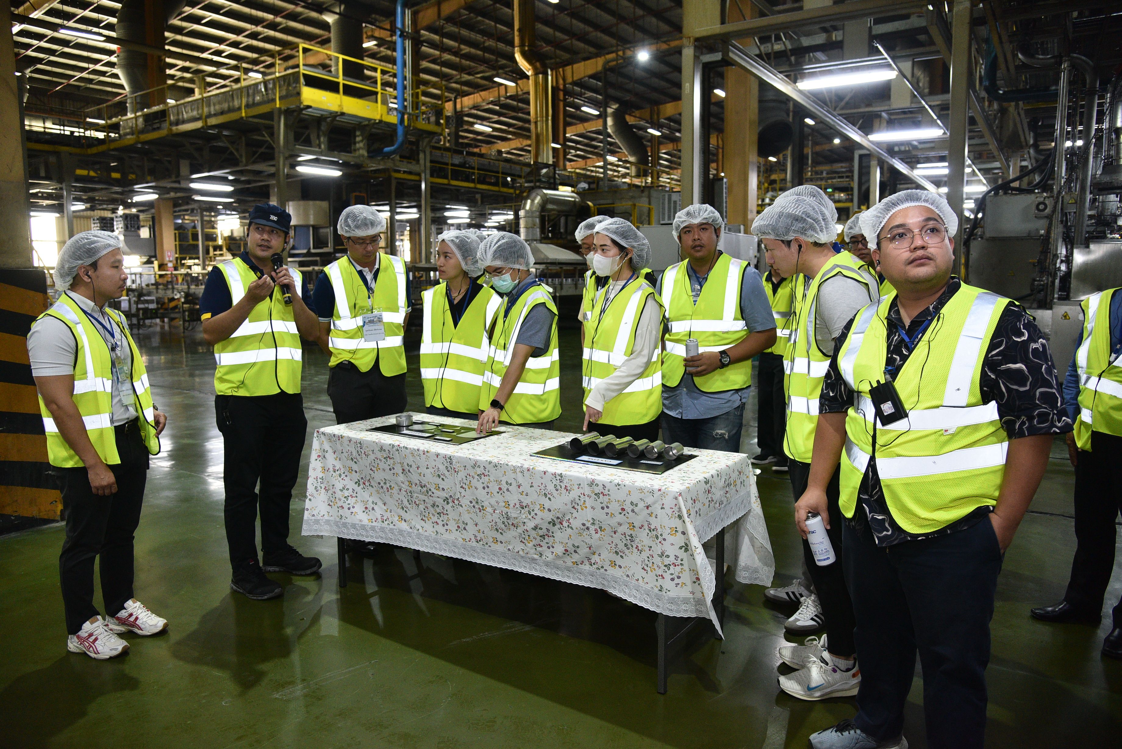 Group in safety vests and hairnets observes products on a table during a factory tour.