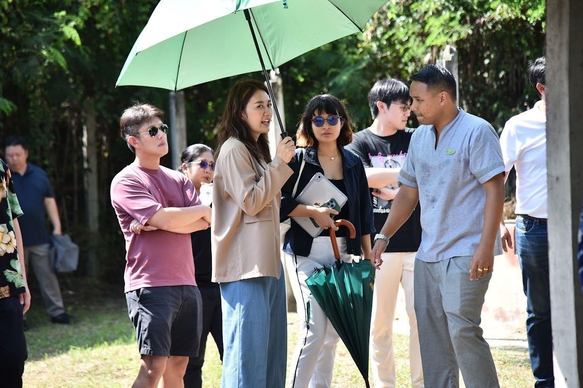 A diverse group of people stands outdoors; one woman holds a green umbrella for shade.