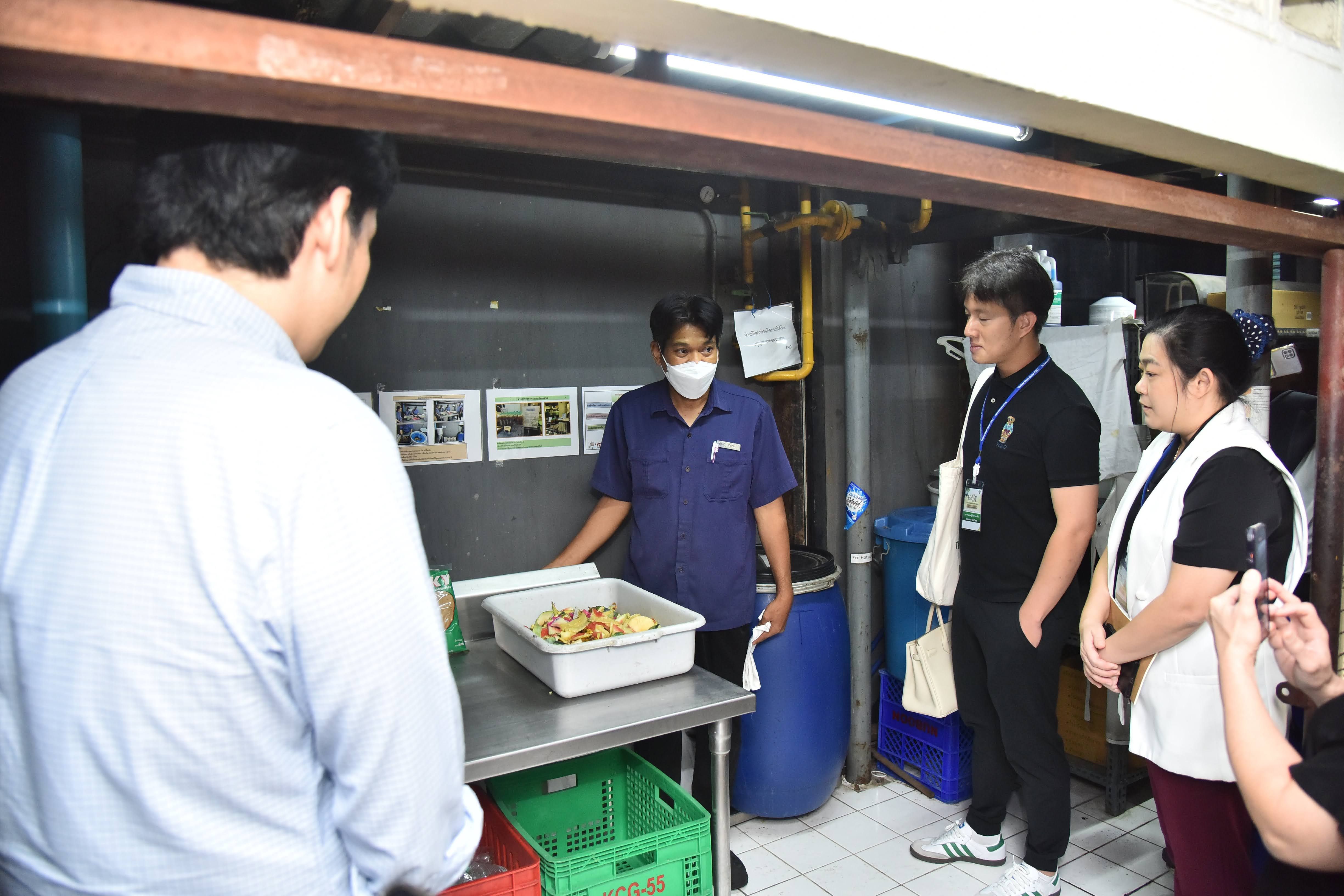 A man in a mask demonstrates food preparation to three people in a commercial kitchen.