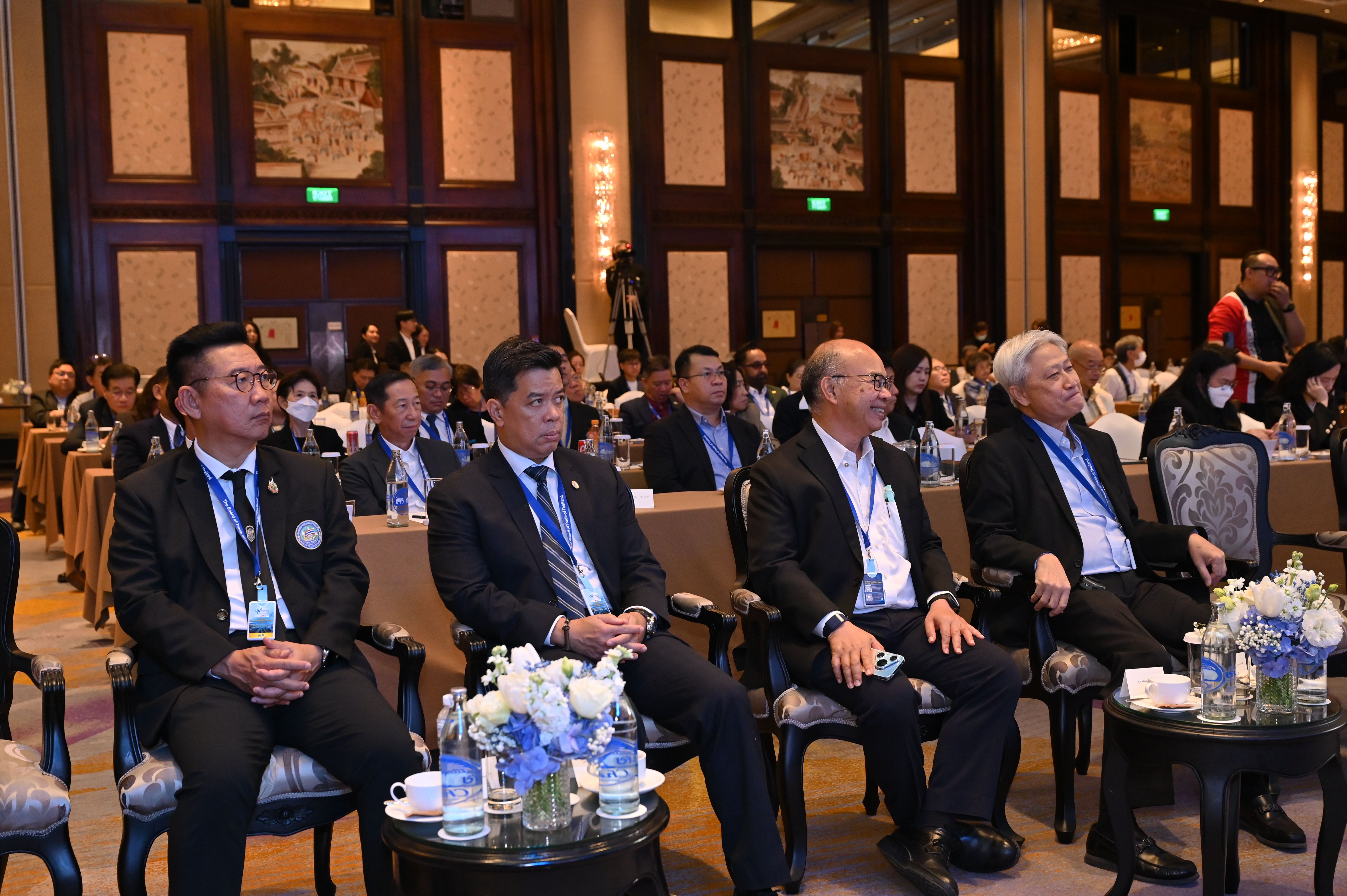 Men in suits attend a conference, seated in a grand hall with elegant decor.