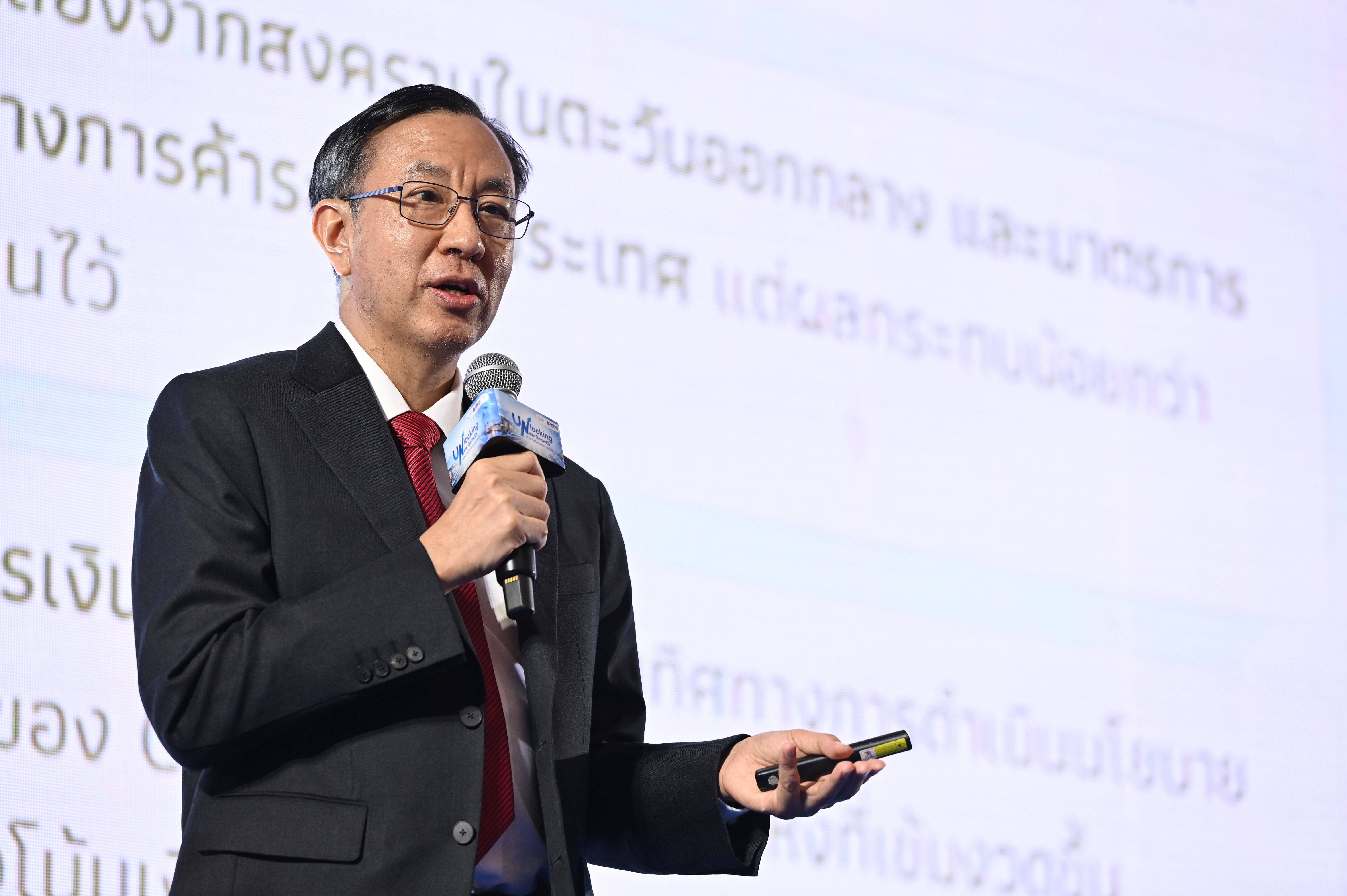 A man in a suit and glasses speaks at a presentation, holding a remote.