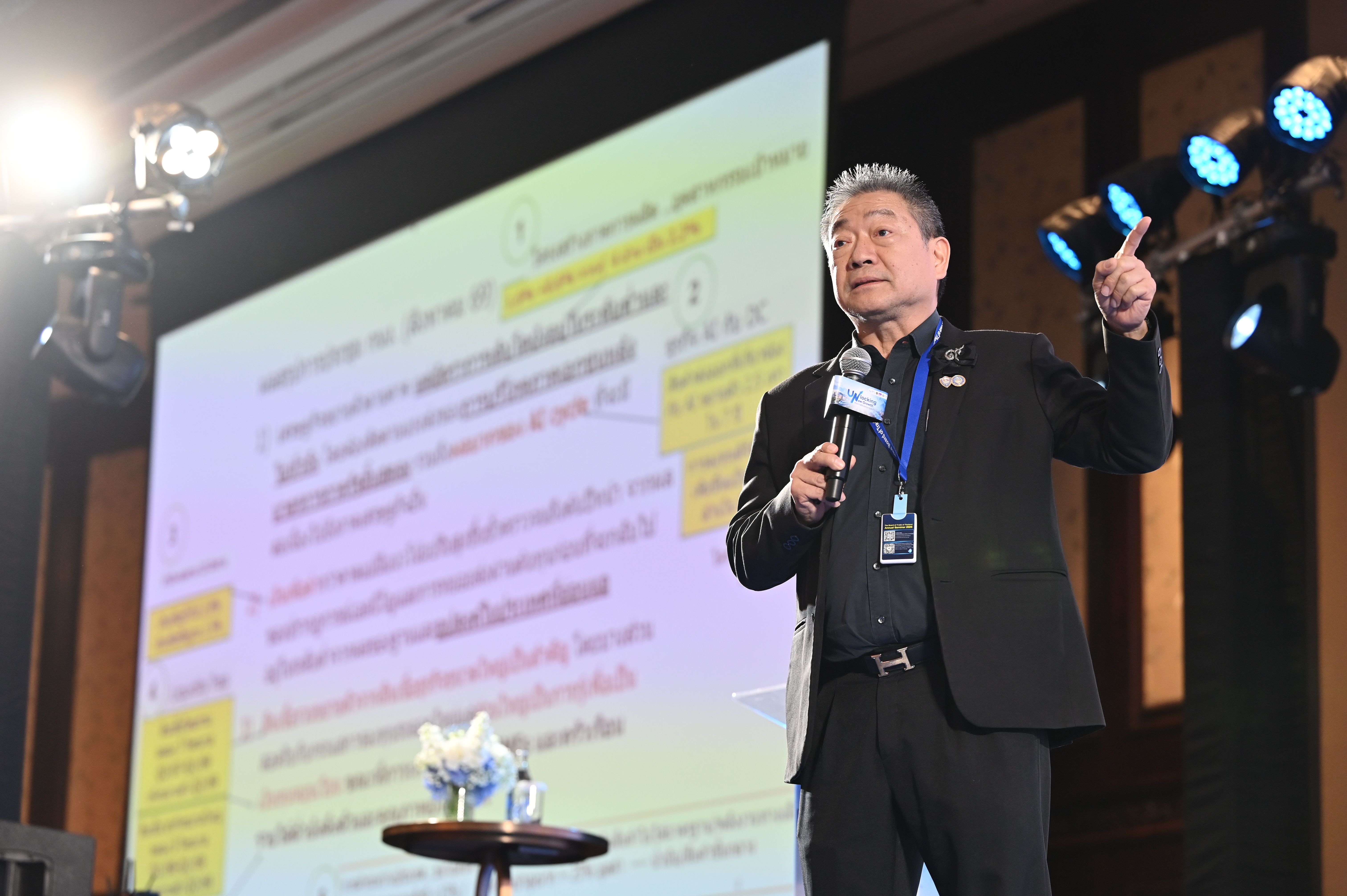 A man in a suit speaks into a microphone, gesturing with a thumbs up.