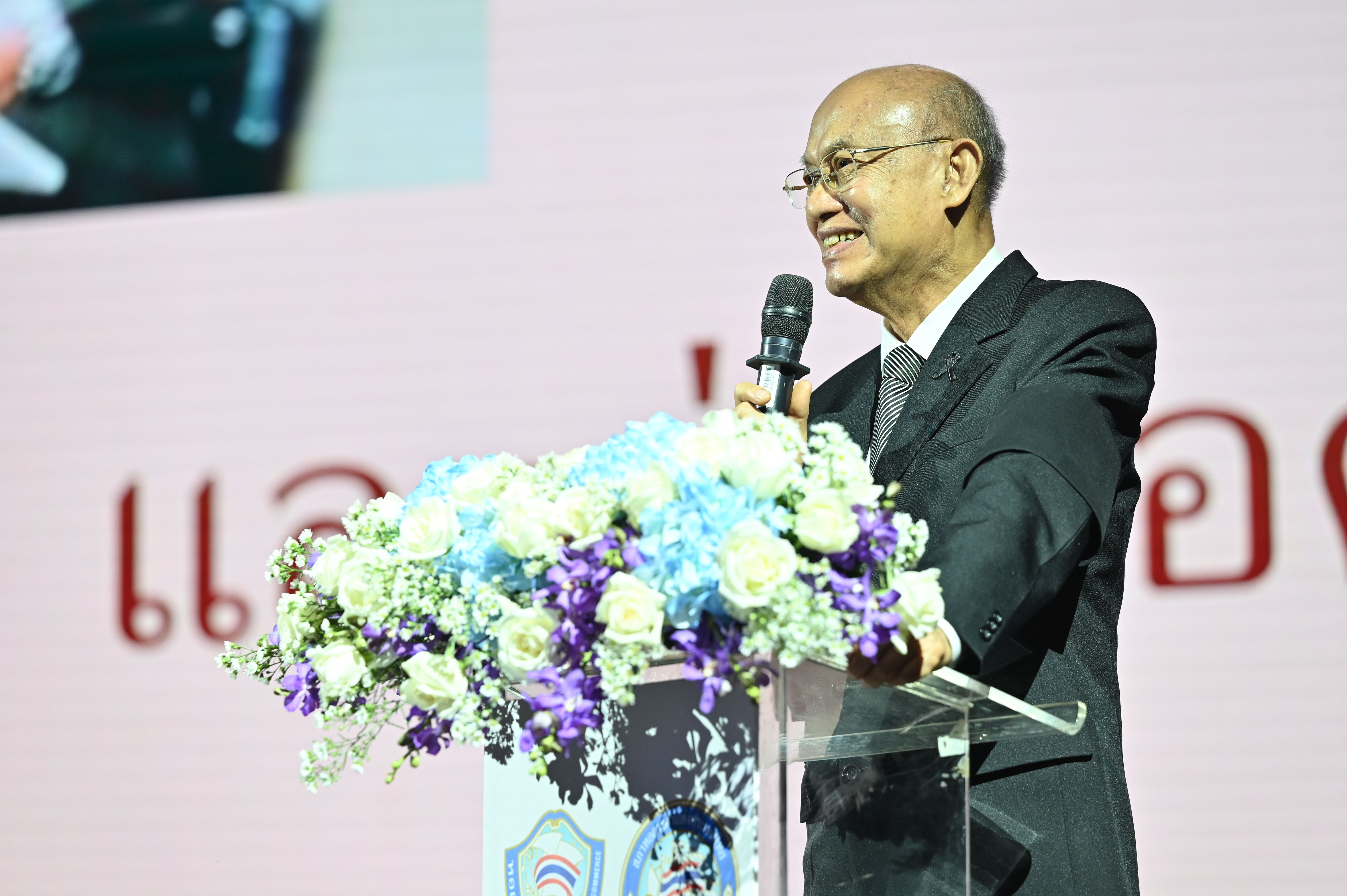 Bald man in a dark suit and glasses smiles while speaking at a podium with flowers.