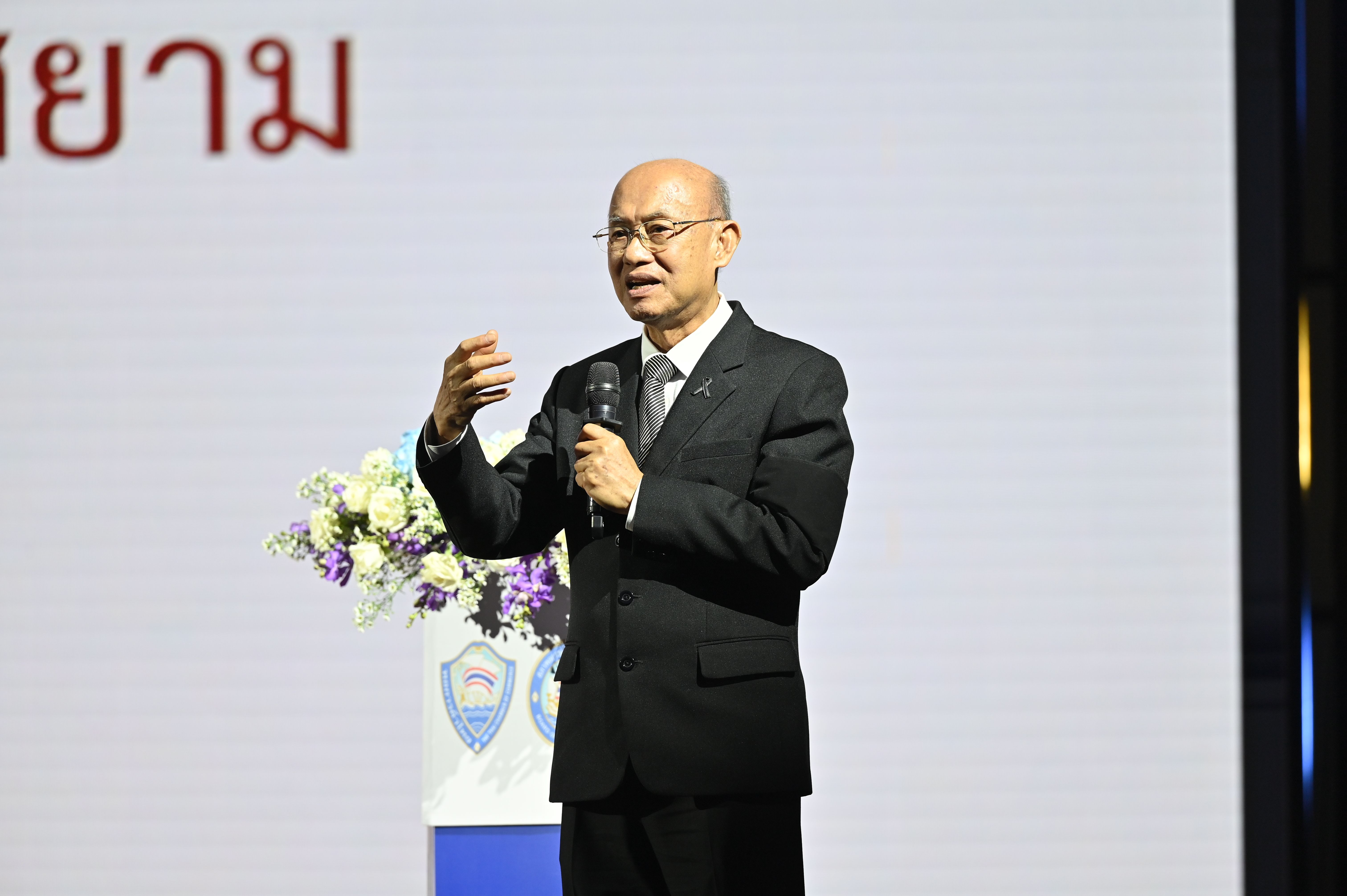 Bald man in glasses and dark suit speaks into microphone, gesturing on stage.