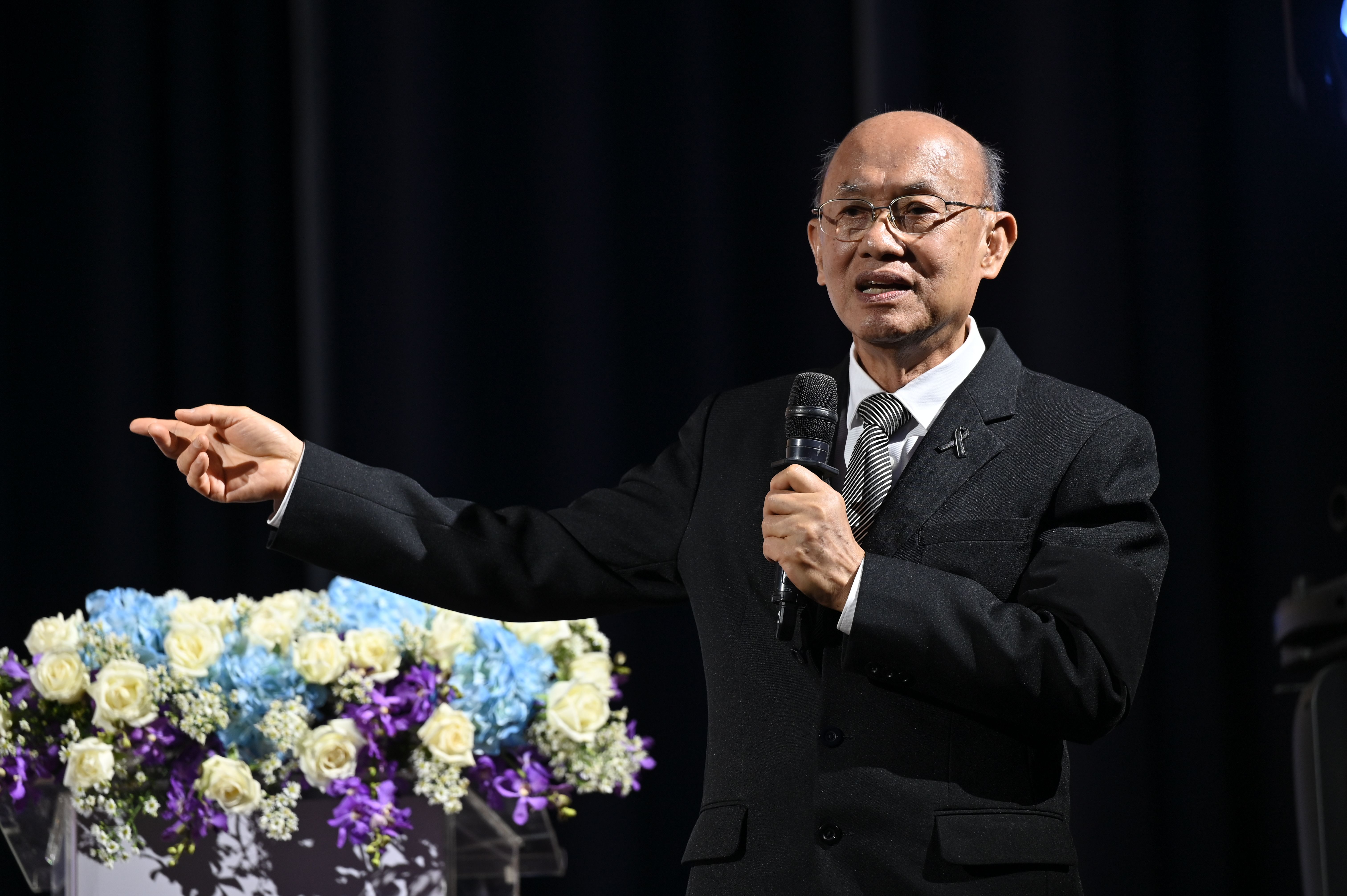 Elderly Asian man with glasses speaks into a microphone, gesturing emphatically.