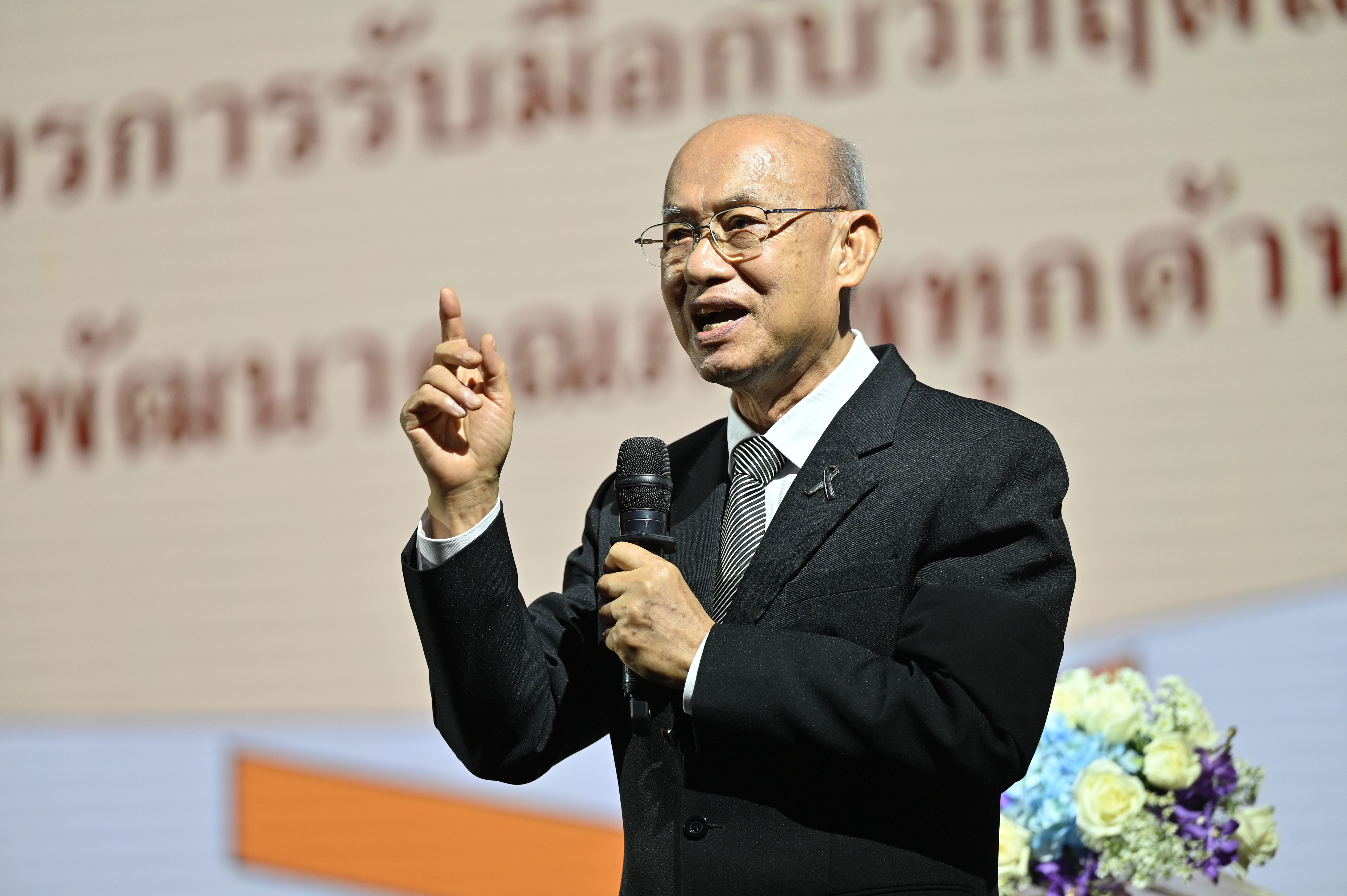 Bald man in suit and glasses speaks into microphone, gesturing during a formal presentation.