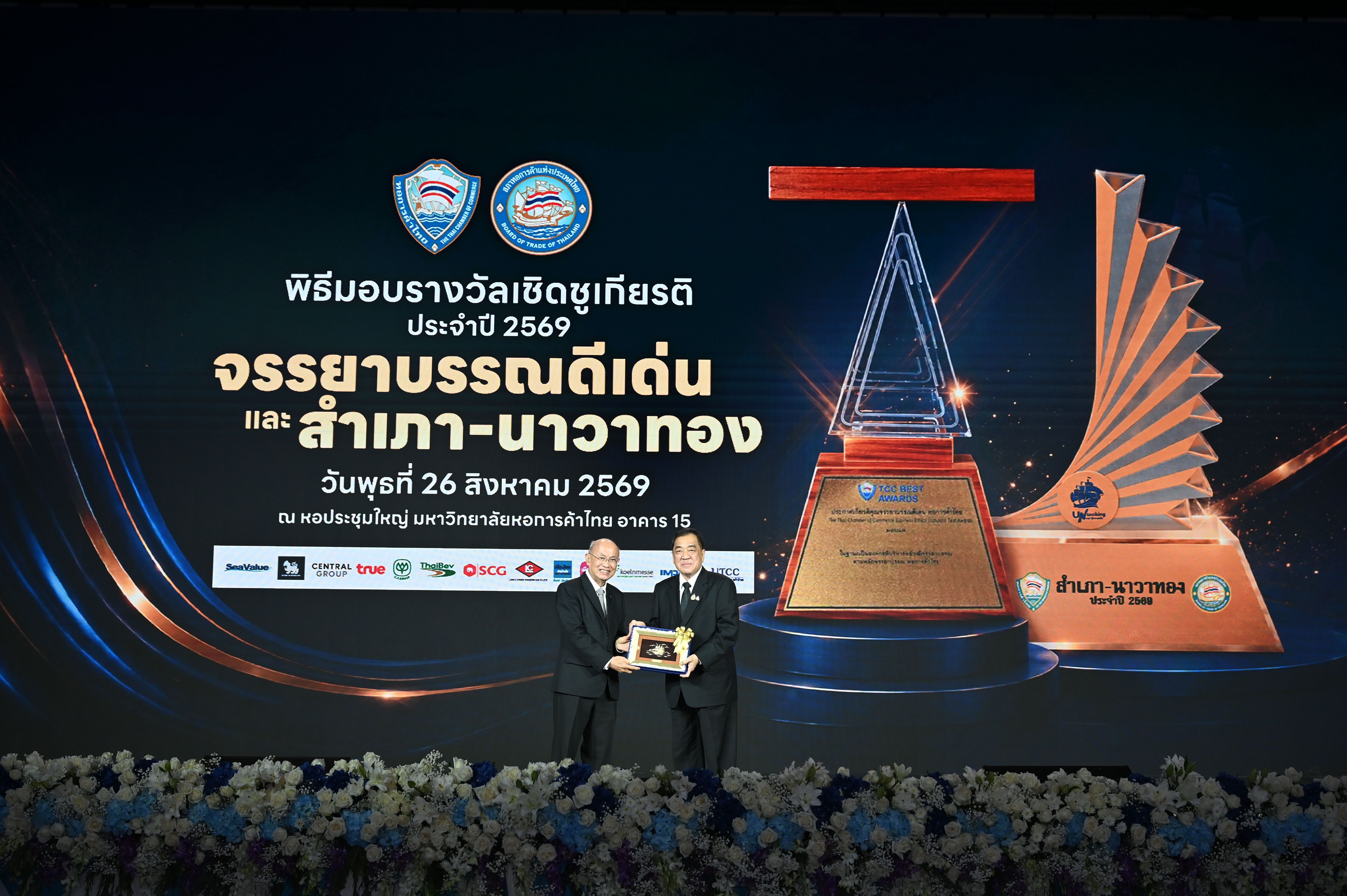 Two men on stage during an award ceremony with large golden trophies displayed.