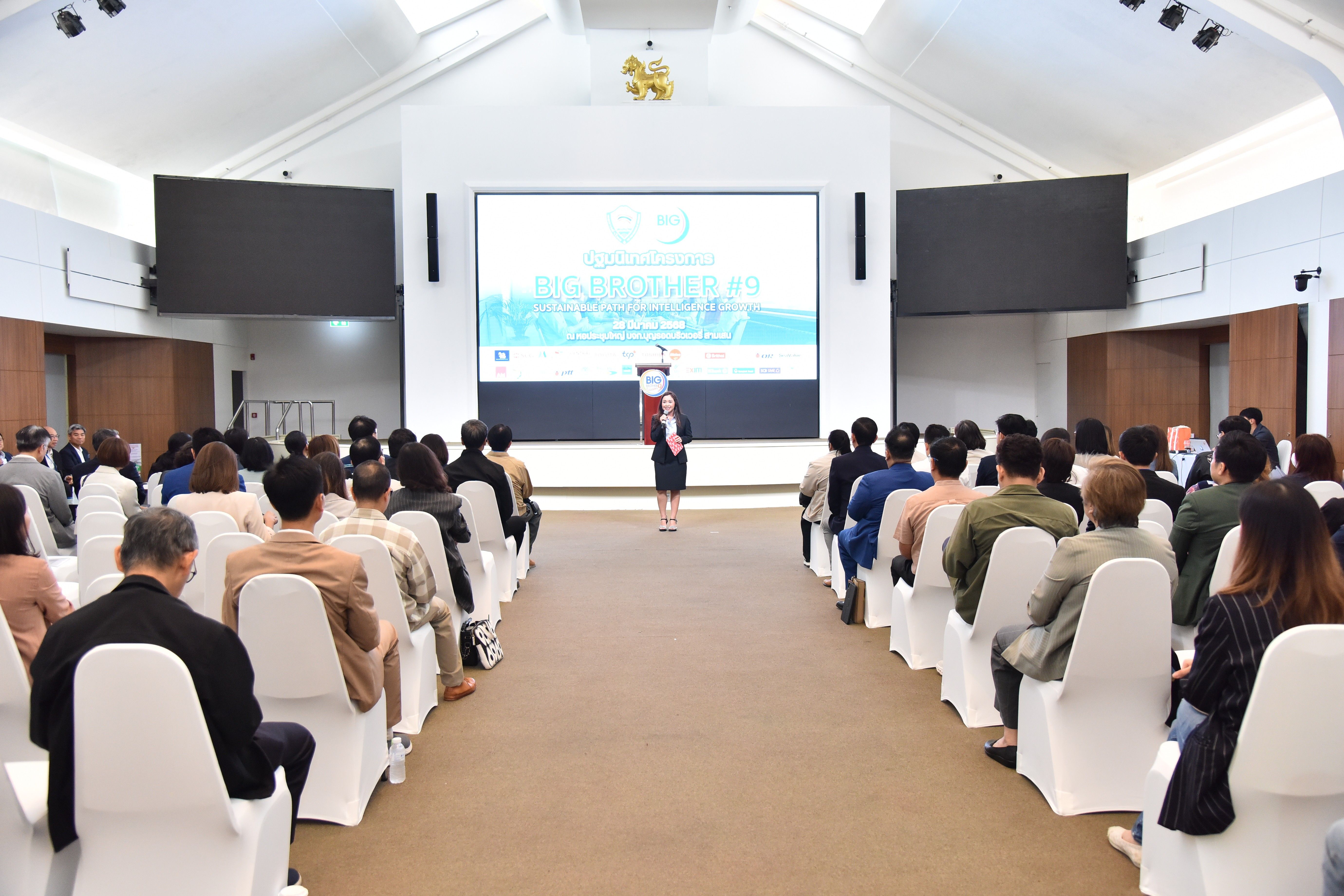 A speaker addresses a seated audience in a modern conference hall with a large projection screen.