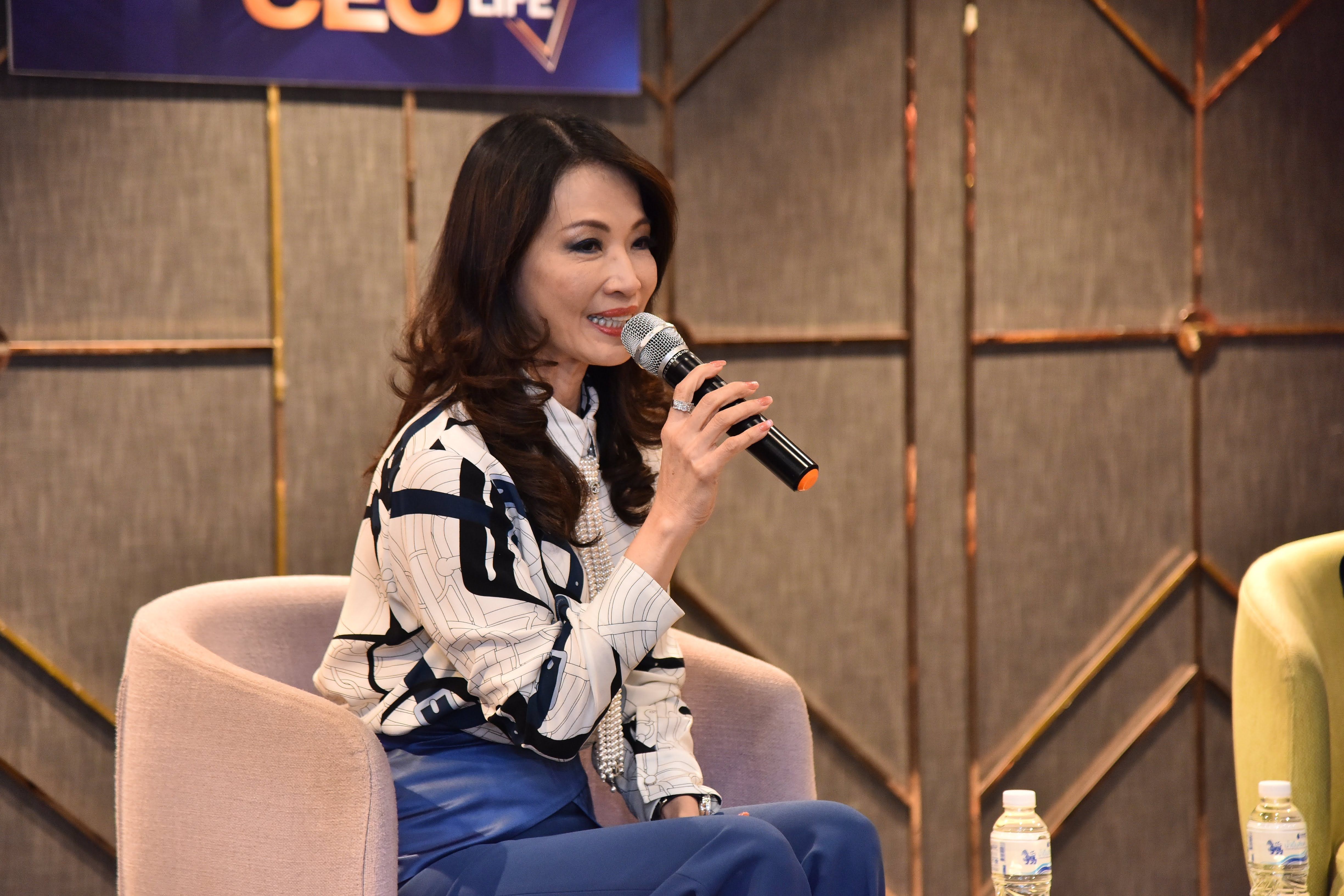 Woman speaking into microphone at an event, seated in a pink armchair.