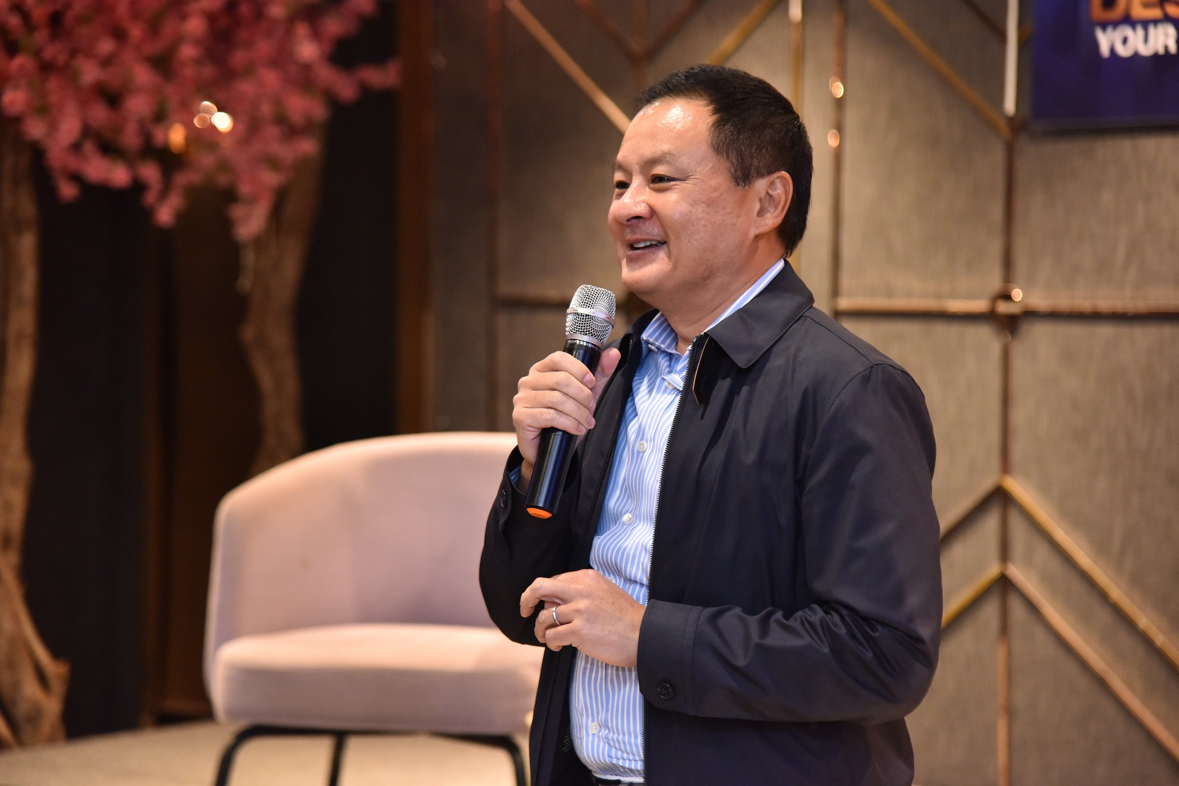 A smiling man in a dark jacket speaks into a microphone at an event.