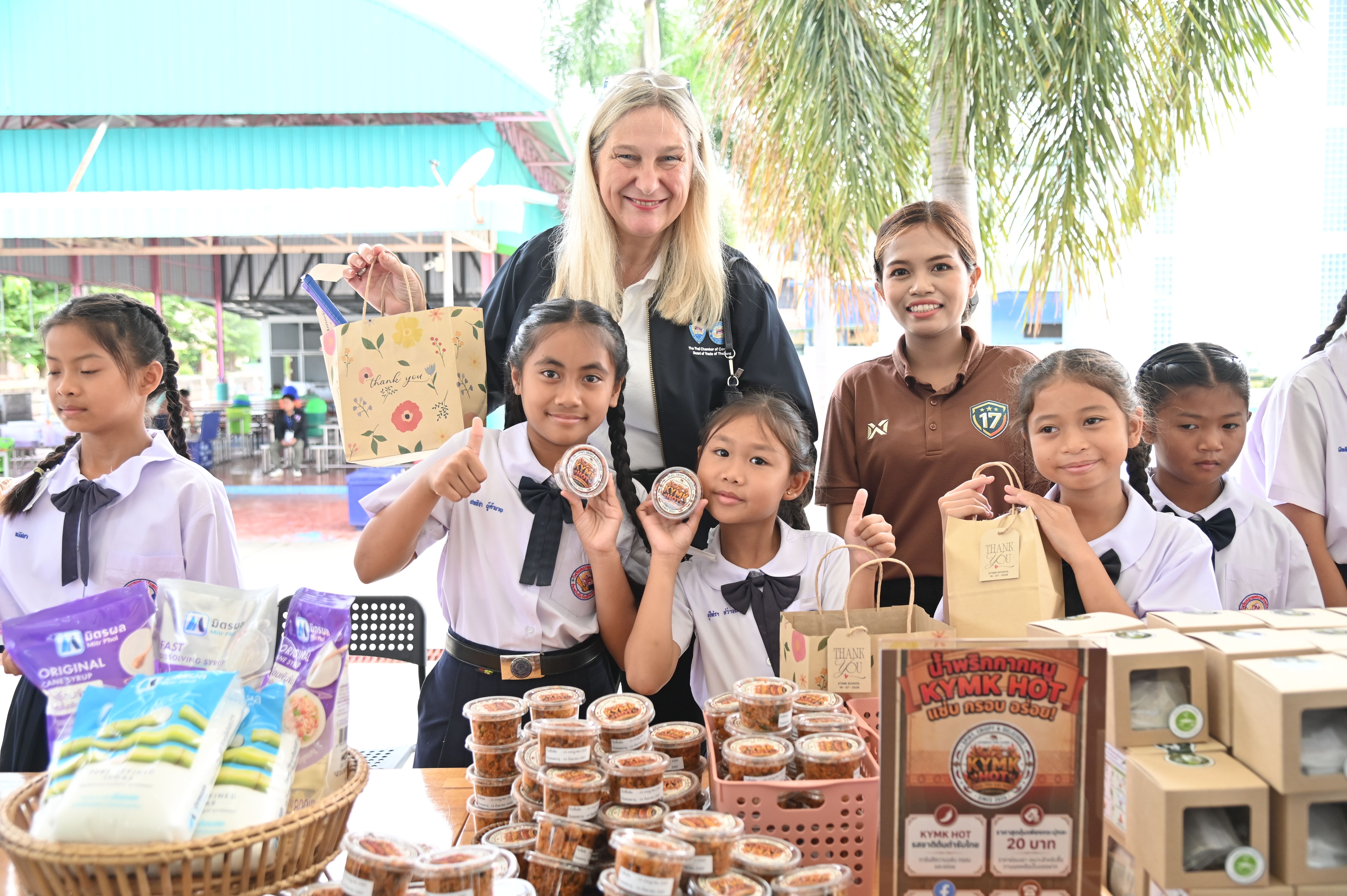 Students and adults proudly display products at a lively outdoor school market stall.