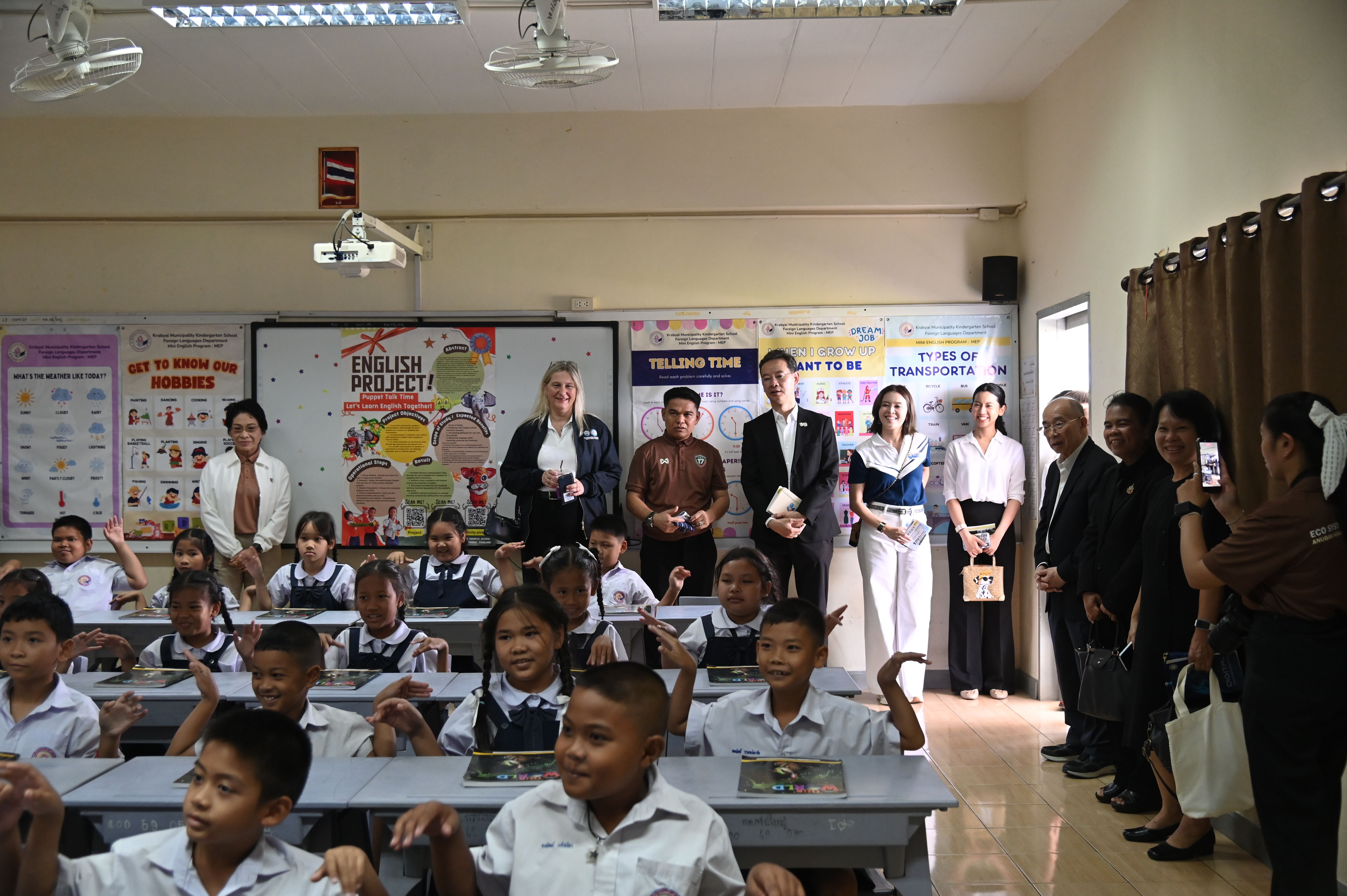 Adults and schoolchildren in a classroom, with some children raising their hands.