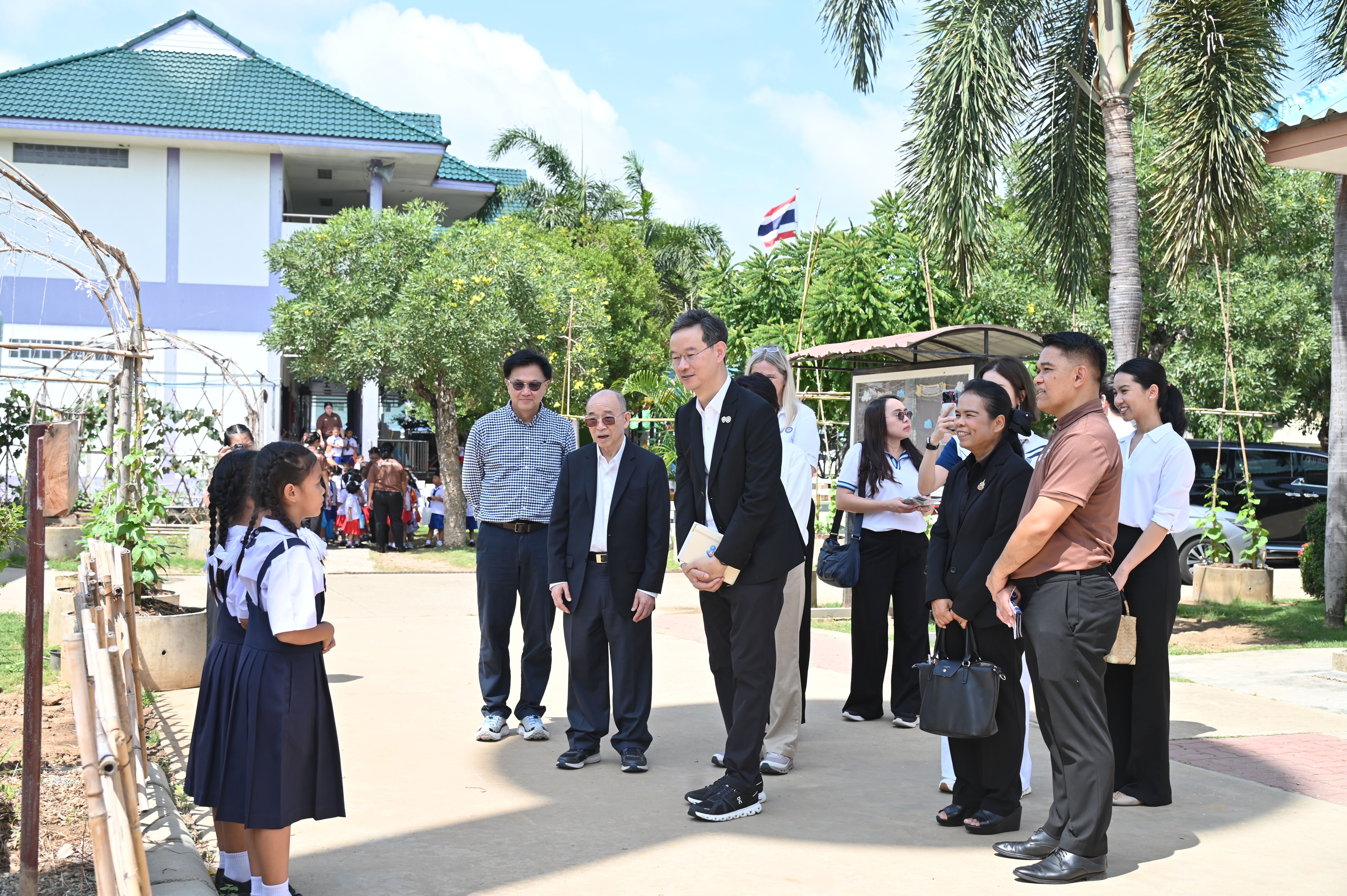 Adults in suits greet young girls in school uniforms outdoors near a school building.
