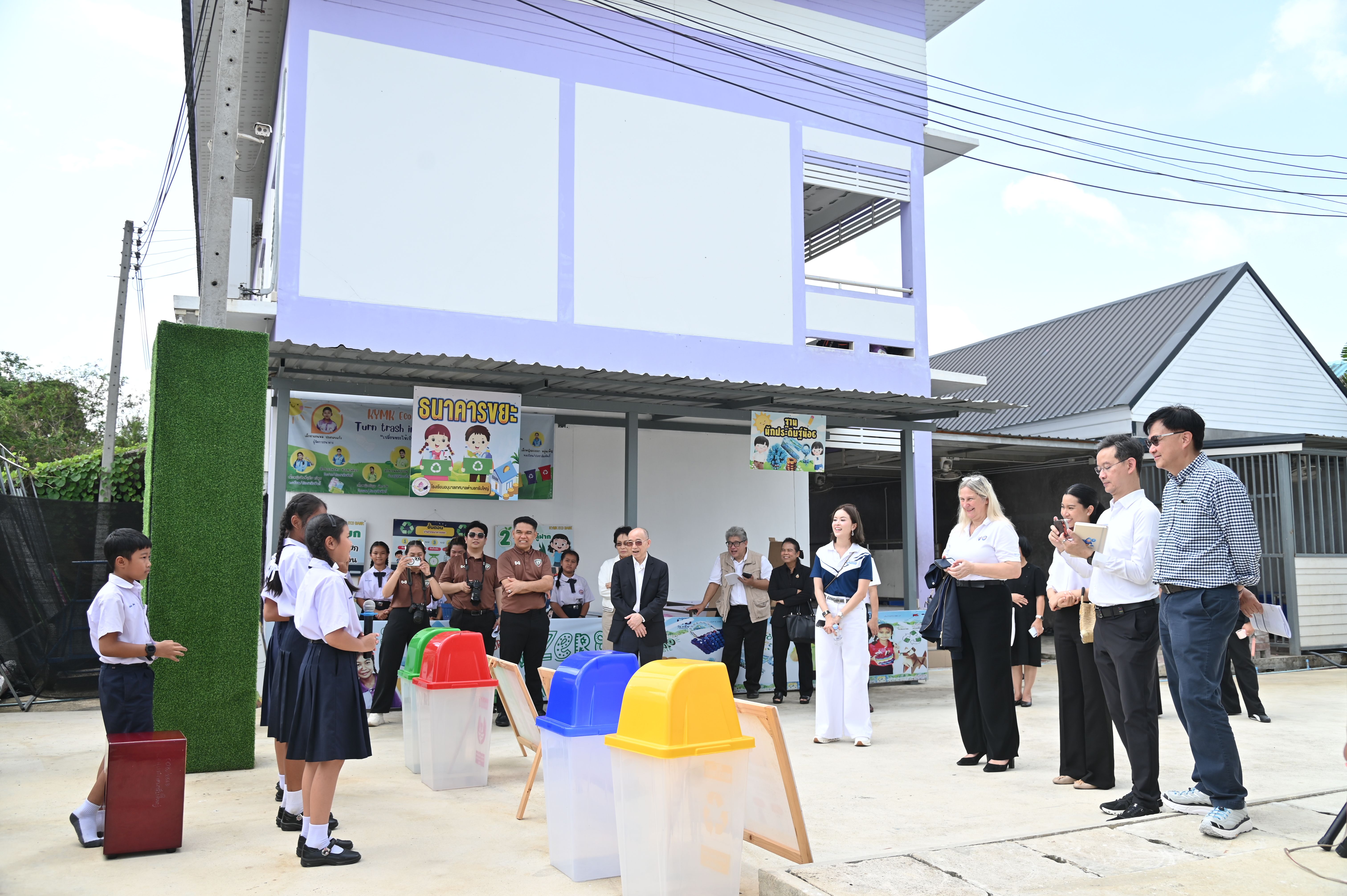 Students and adults gather outside a building with colorful recycling bins.