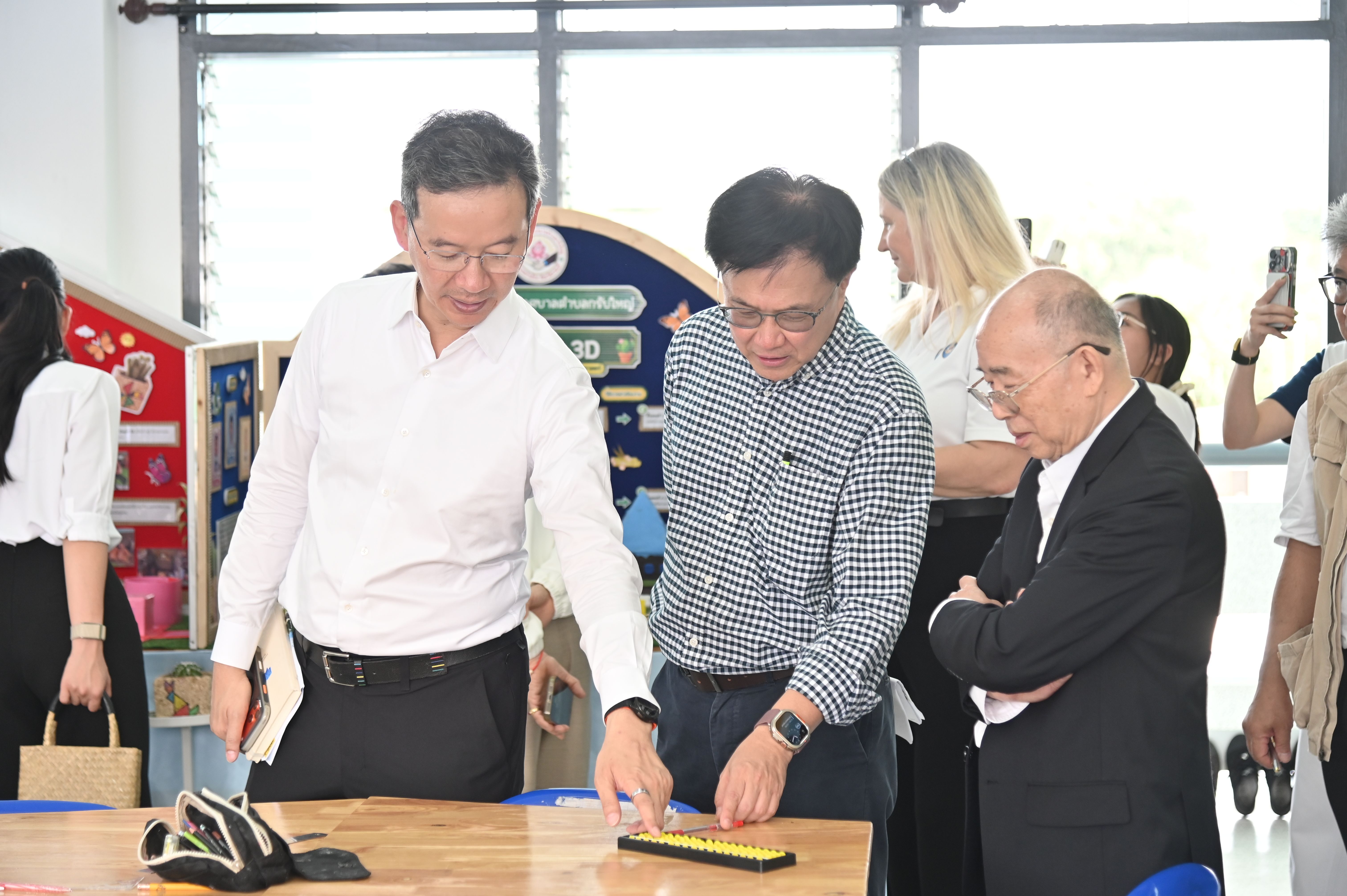 Two men intently observe an object on a wooden table with others nearby.