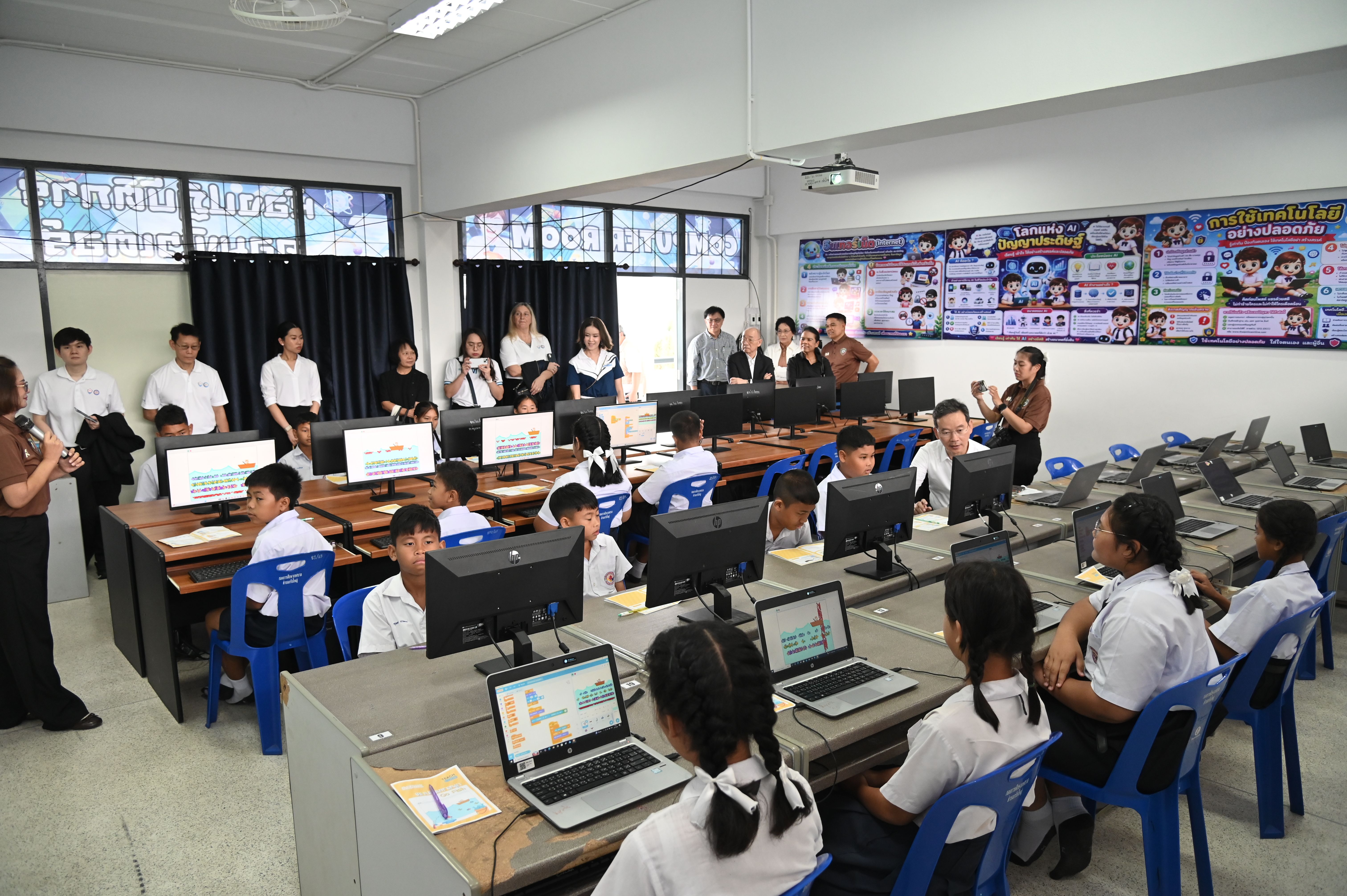 Students in uniforms learn on computers in a modern classroom, observed by several adults.