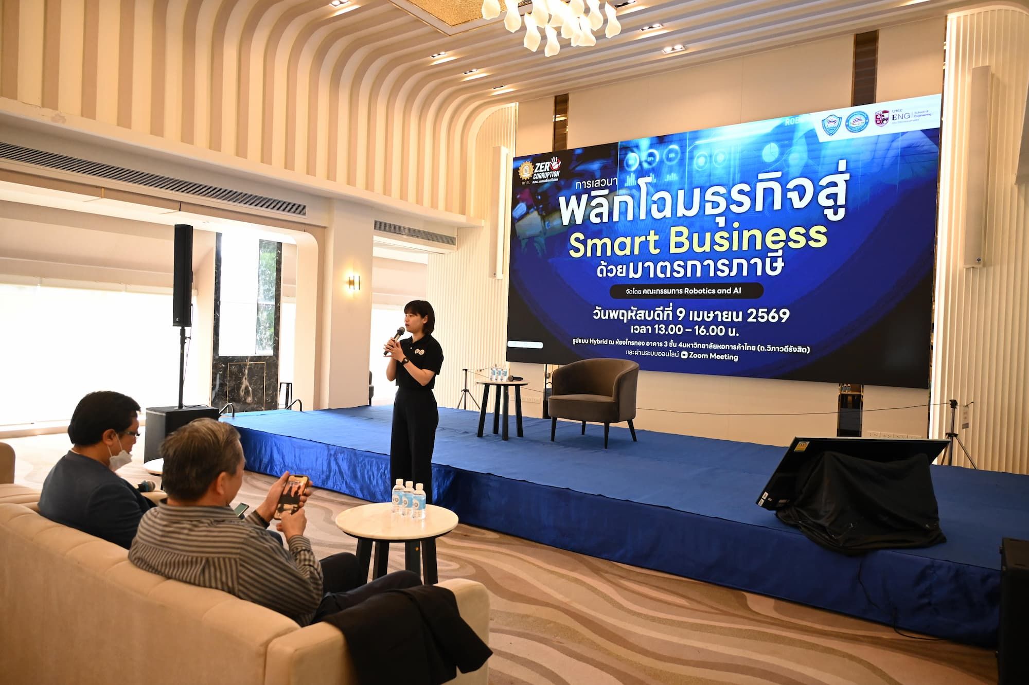 Woman presenter on stage speaking to an audience with a large screen displaying 'Smart Business'.