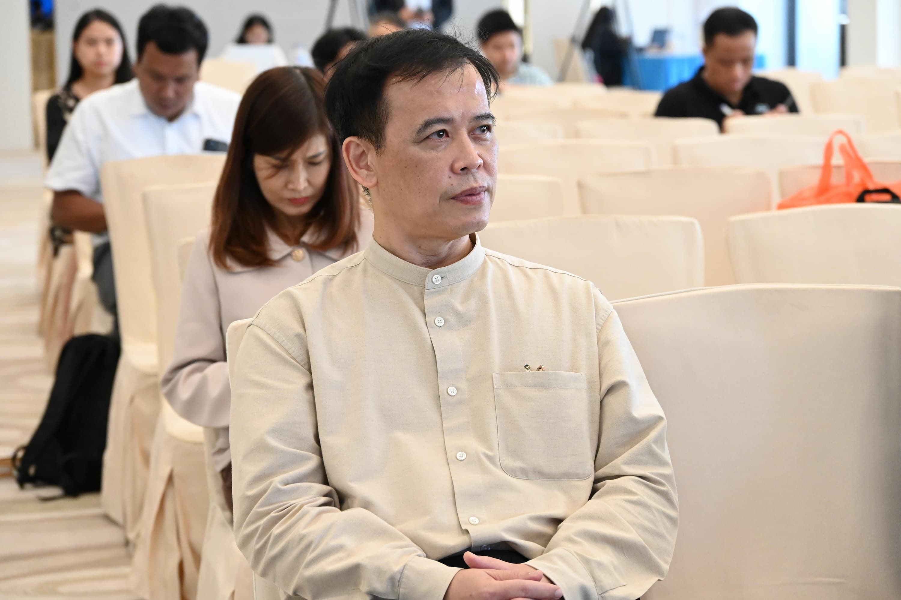 Man in light beige shirt sits in audience, looking thoughtfully right.