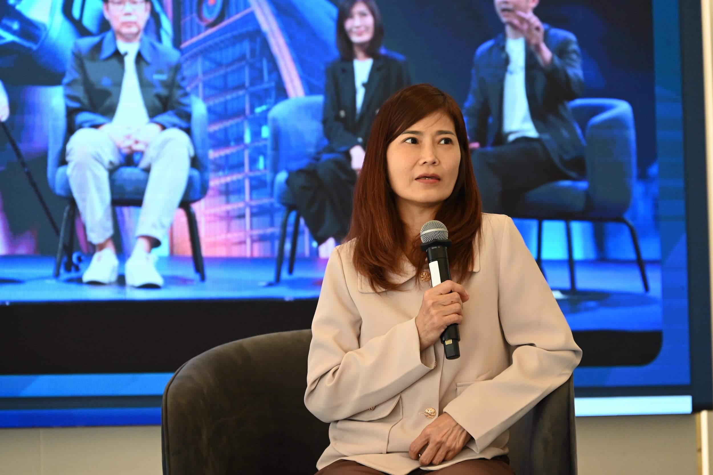 A woman with long brown hair speaks into a microphone while seated on a stage.
