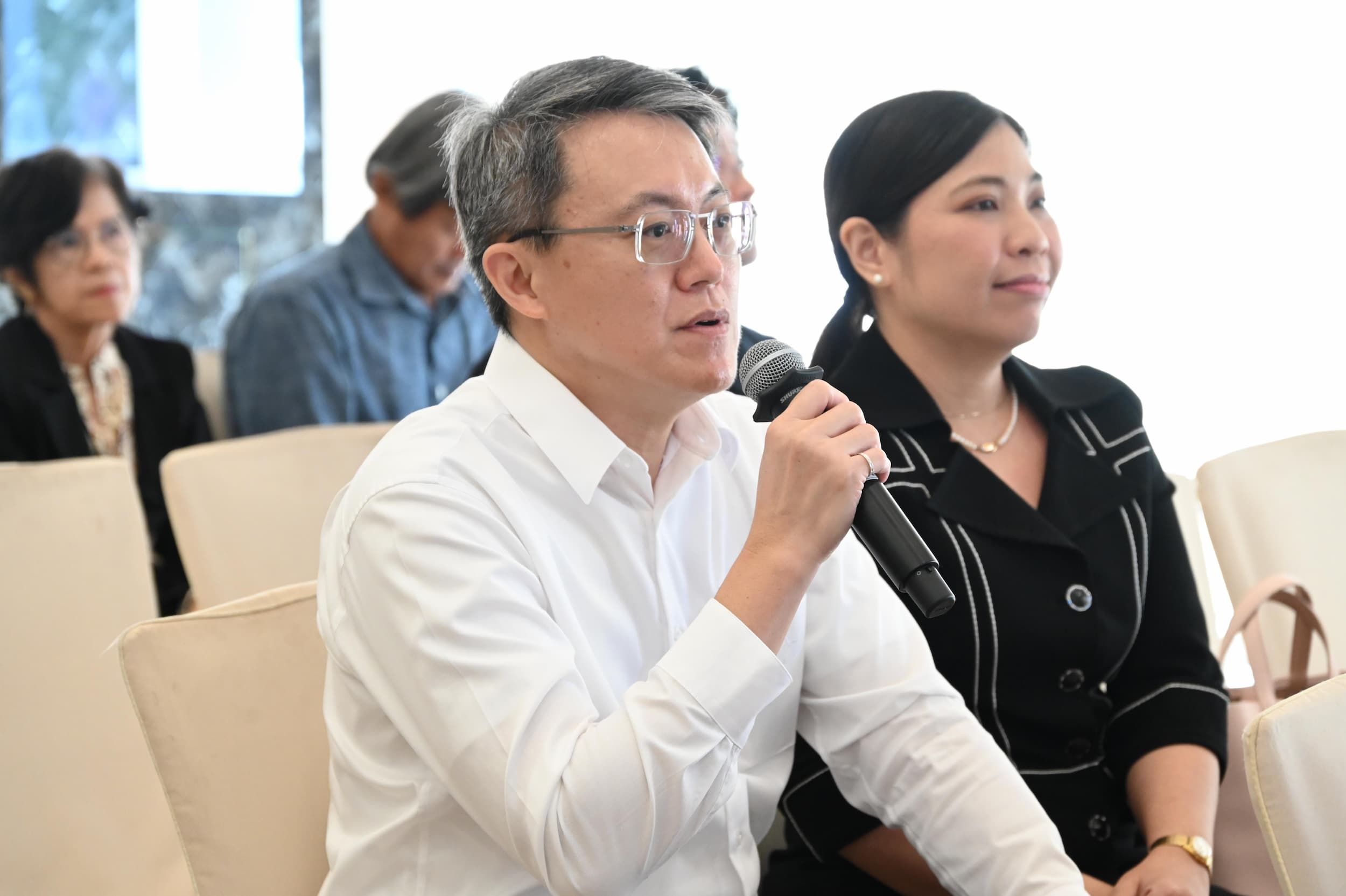Man in white shirt and glasses speaking into a microphone at a professional event.