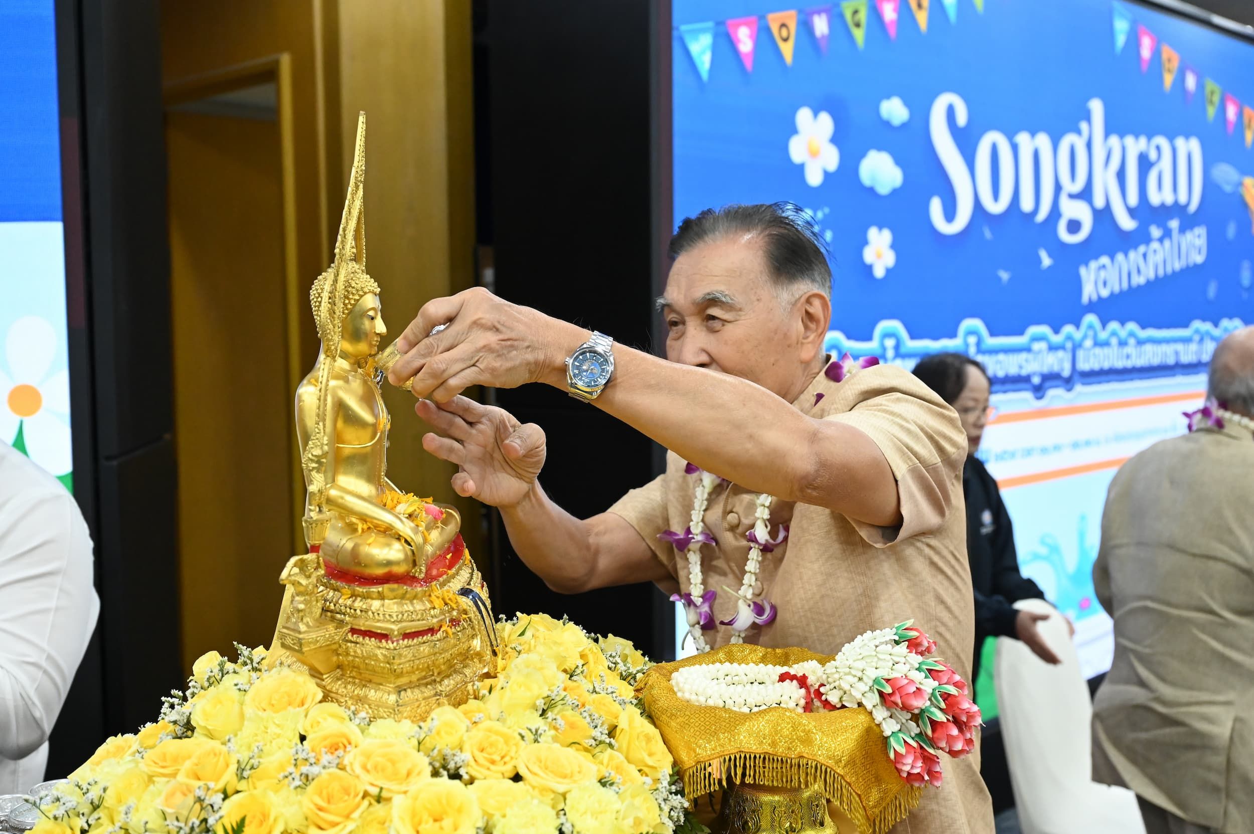 Older man in brown pours water over a golden Buddha statue decorated with yellow flowers, celebrating Songkran.
