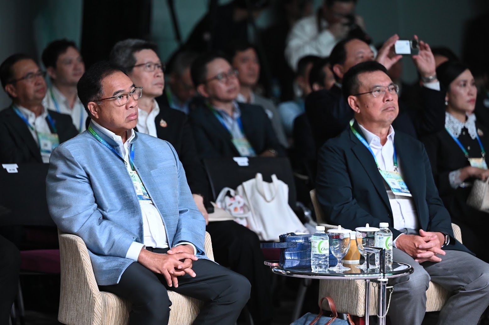 Two men in business attire sit attentively in an audience at a conference.