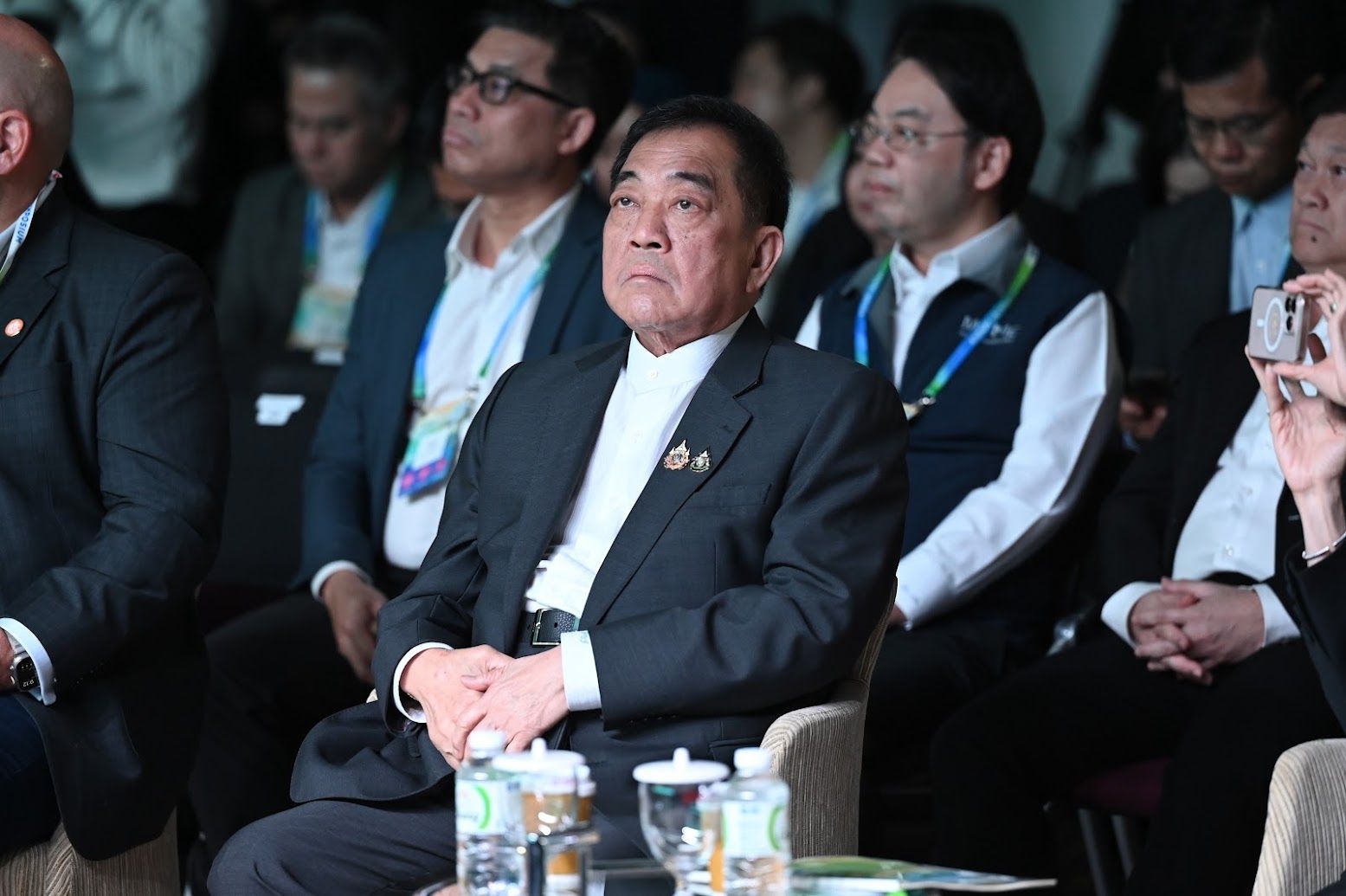 Formal Asian man in a suit attentively observing an event, hands clasped, serious expression.
