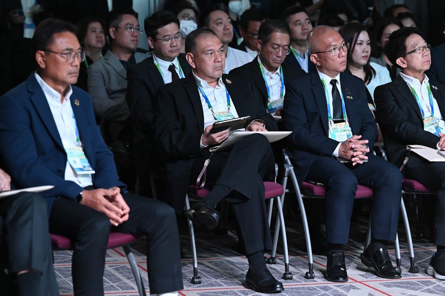 Men in business attire sit attentively in a conference audience, listening to a presentation.