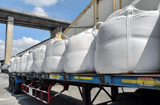 A truck trailer loaded with many large white industrial bags under a concrete bridge.