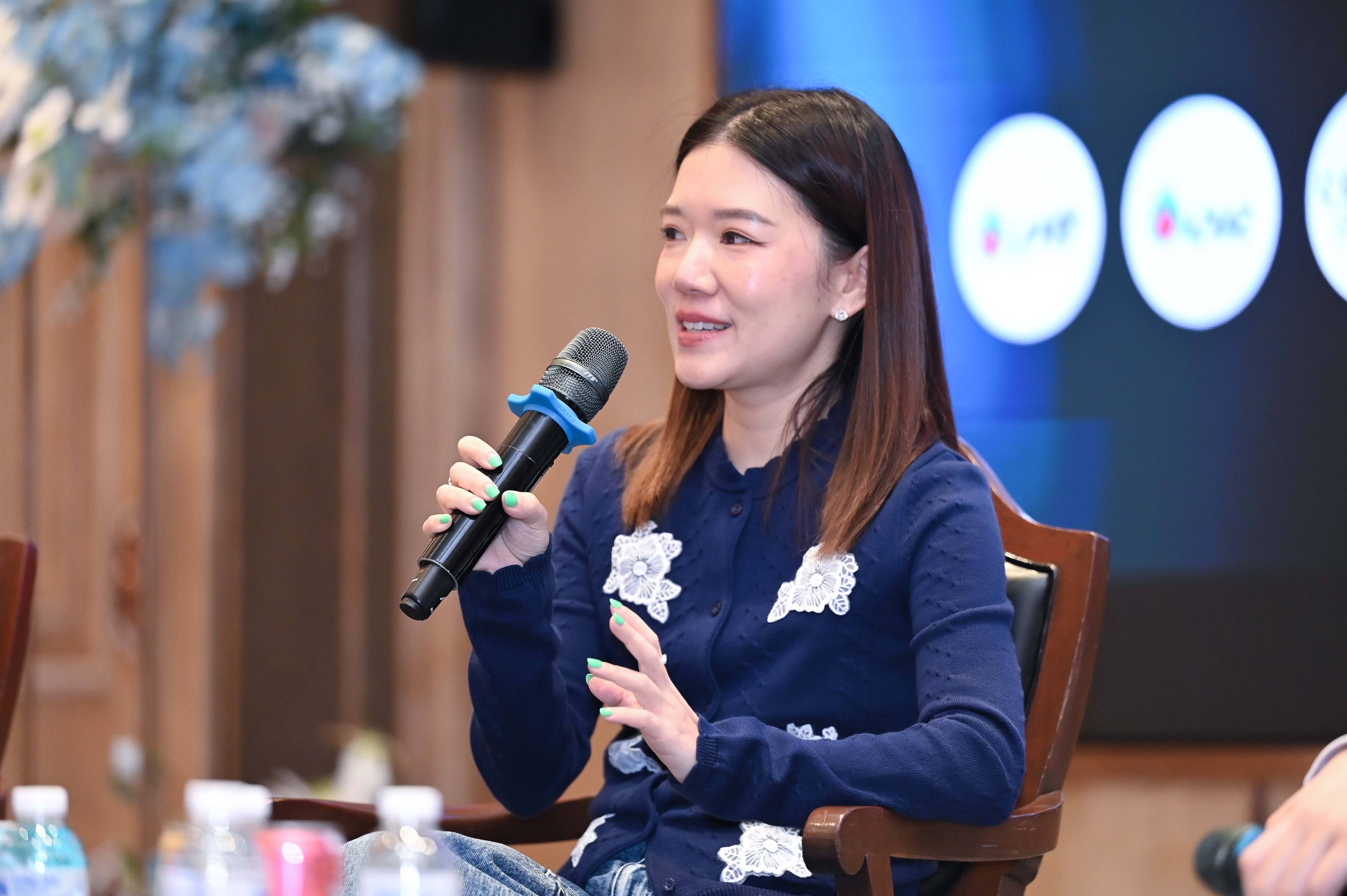 A woman speaking into a microphone at a conference, gesturing with her hand.