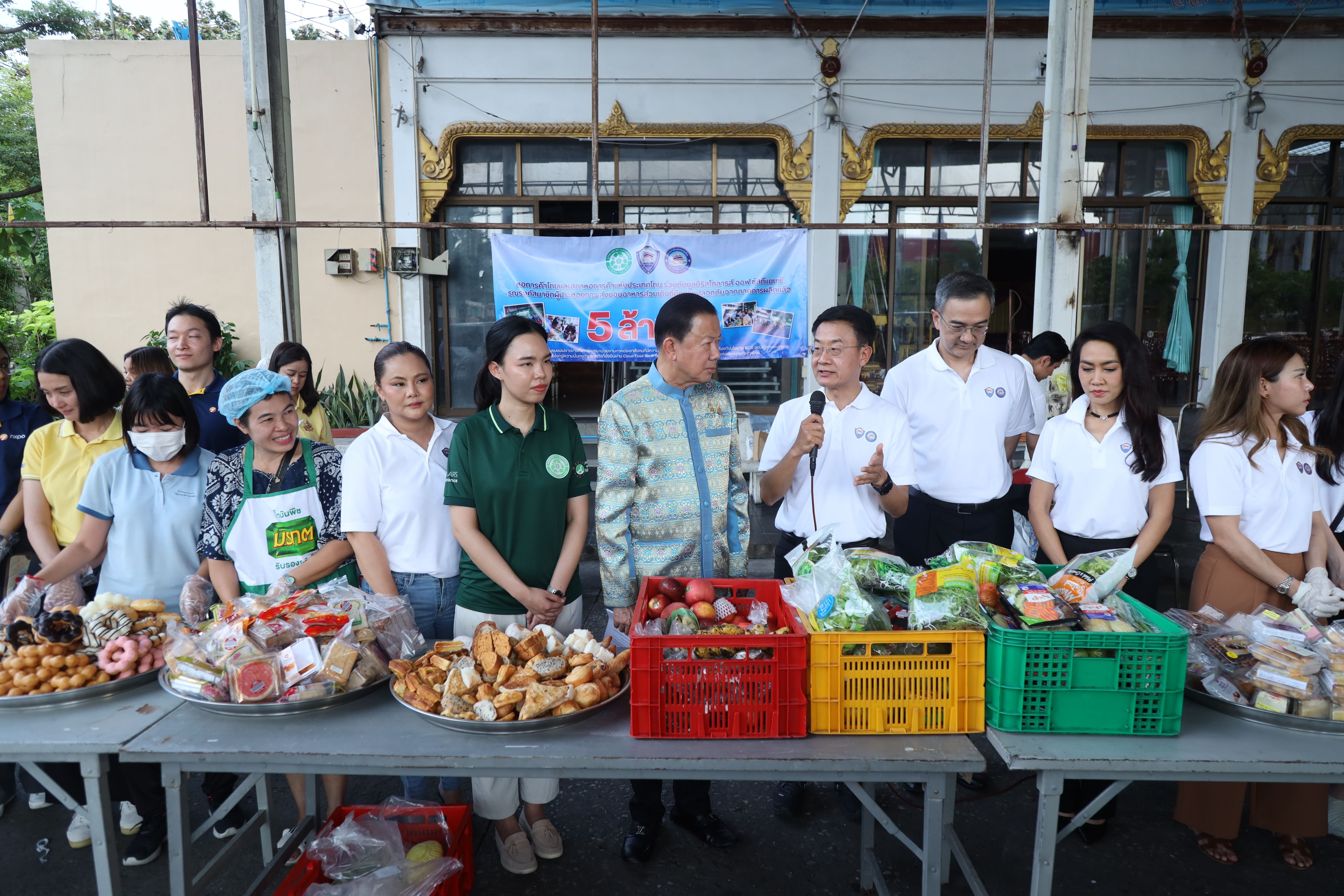 Group of people and a man speaking behind tables with food donations at a community event.