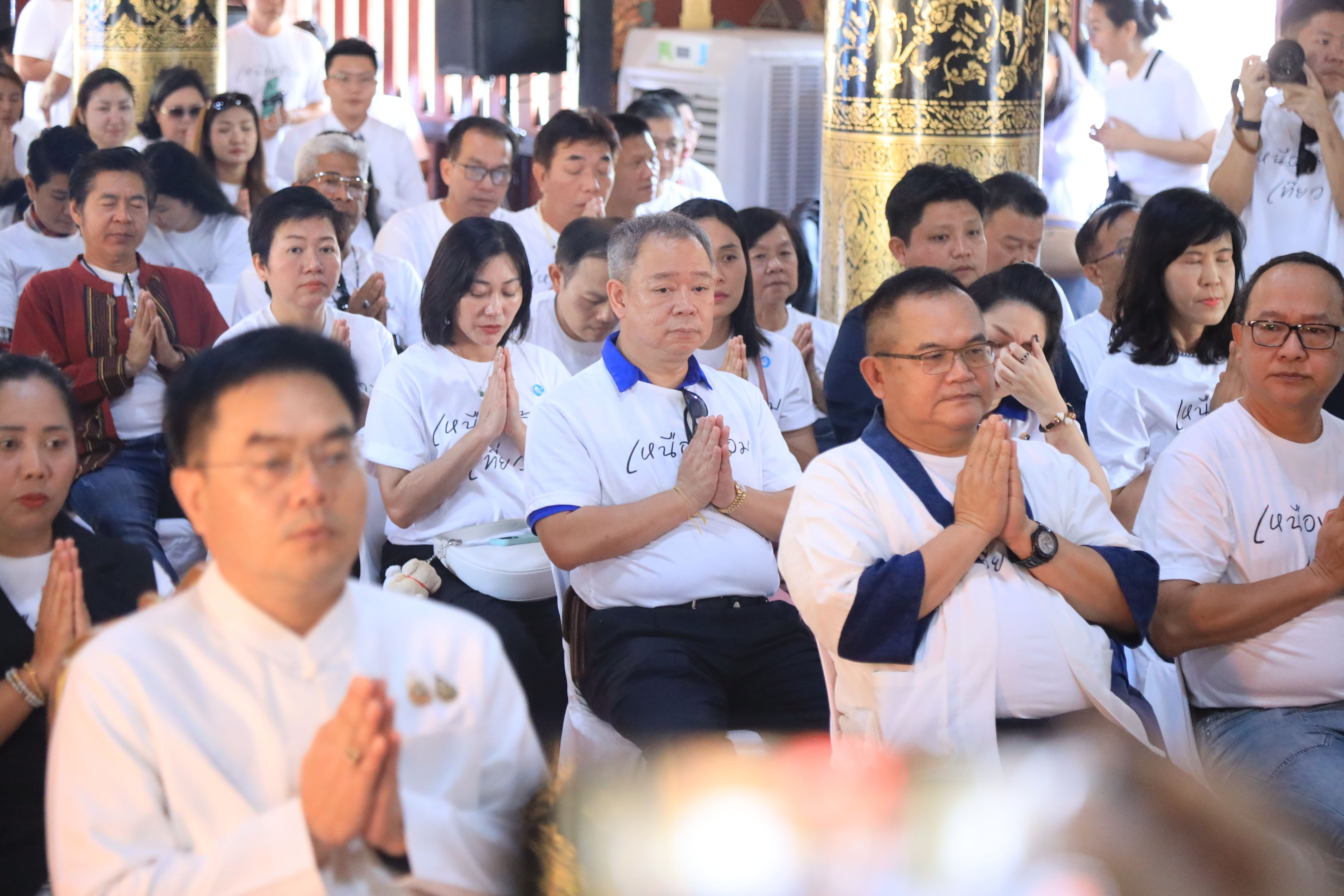Attendees in white shirts sitting with hands in prayer during a spiritual ceremony.