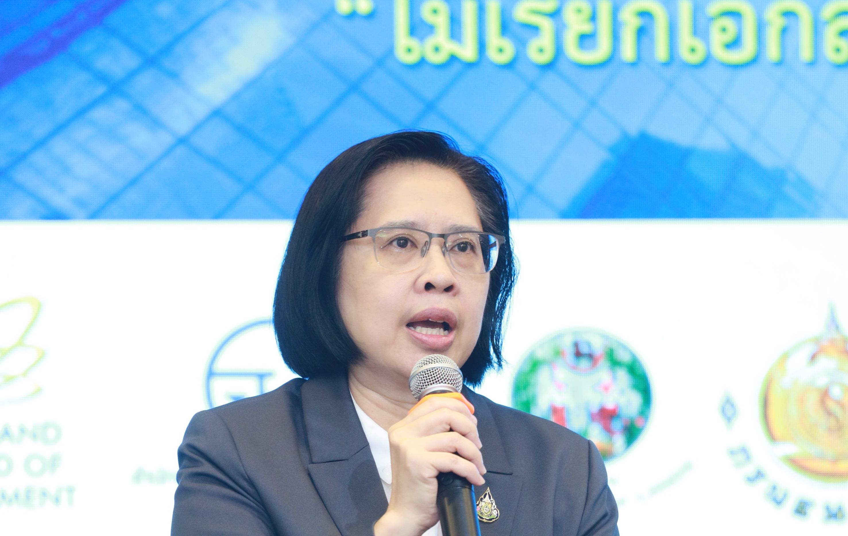 A woman with glasses and dark suit jacket speaks into a microphone at an event.