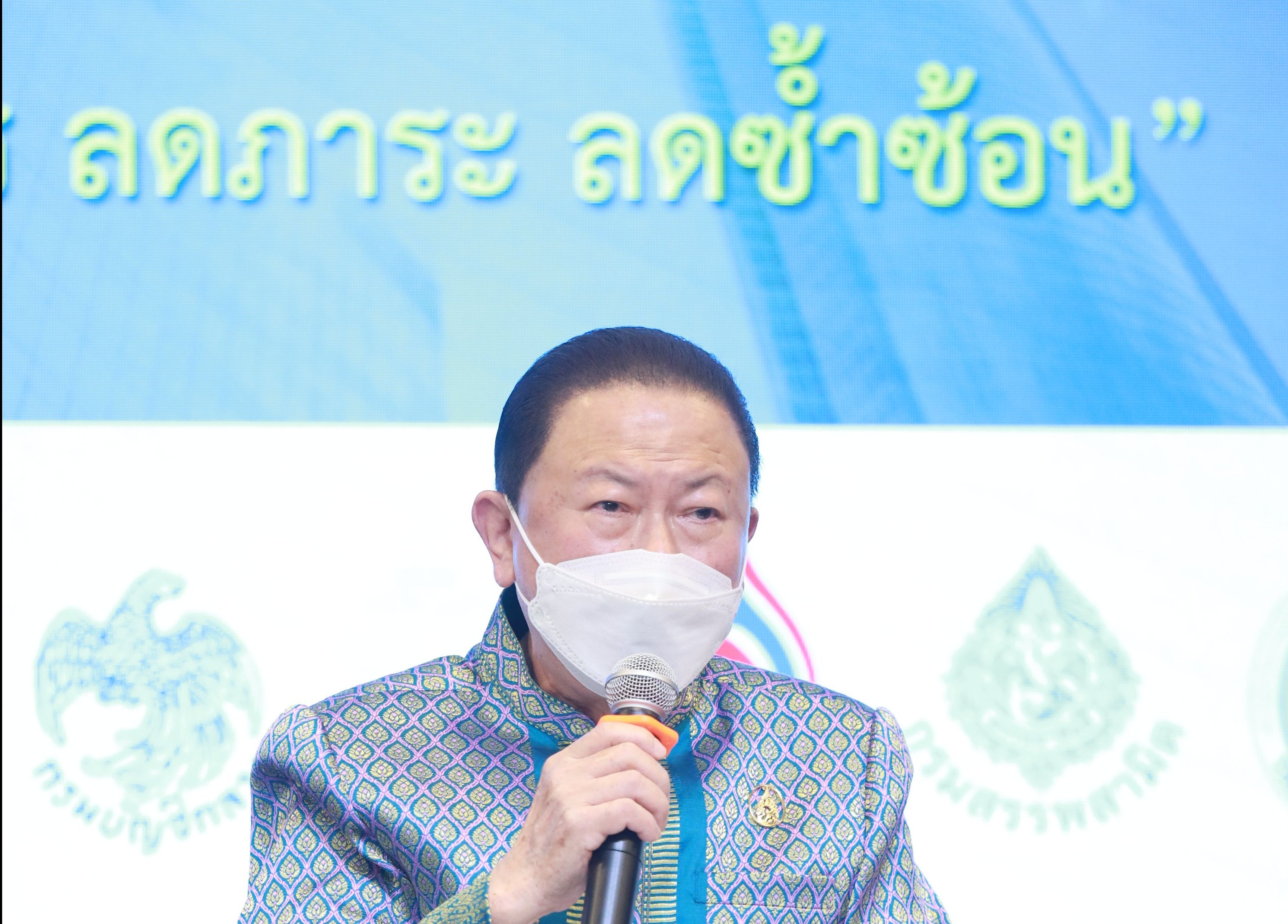 Man speaking into microphone while wearing a traditional patterned shirt and white face mask.