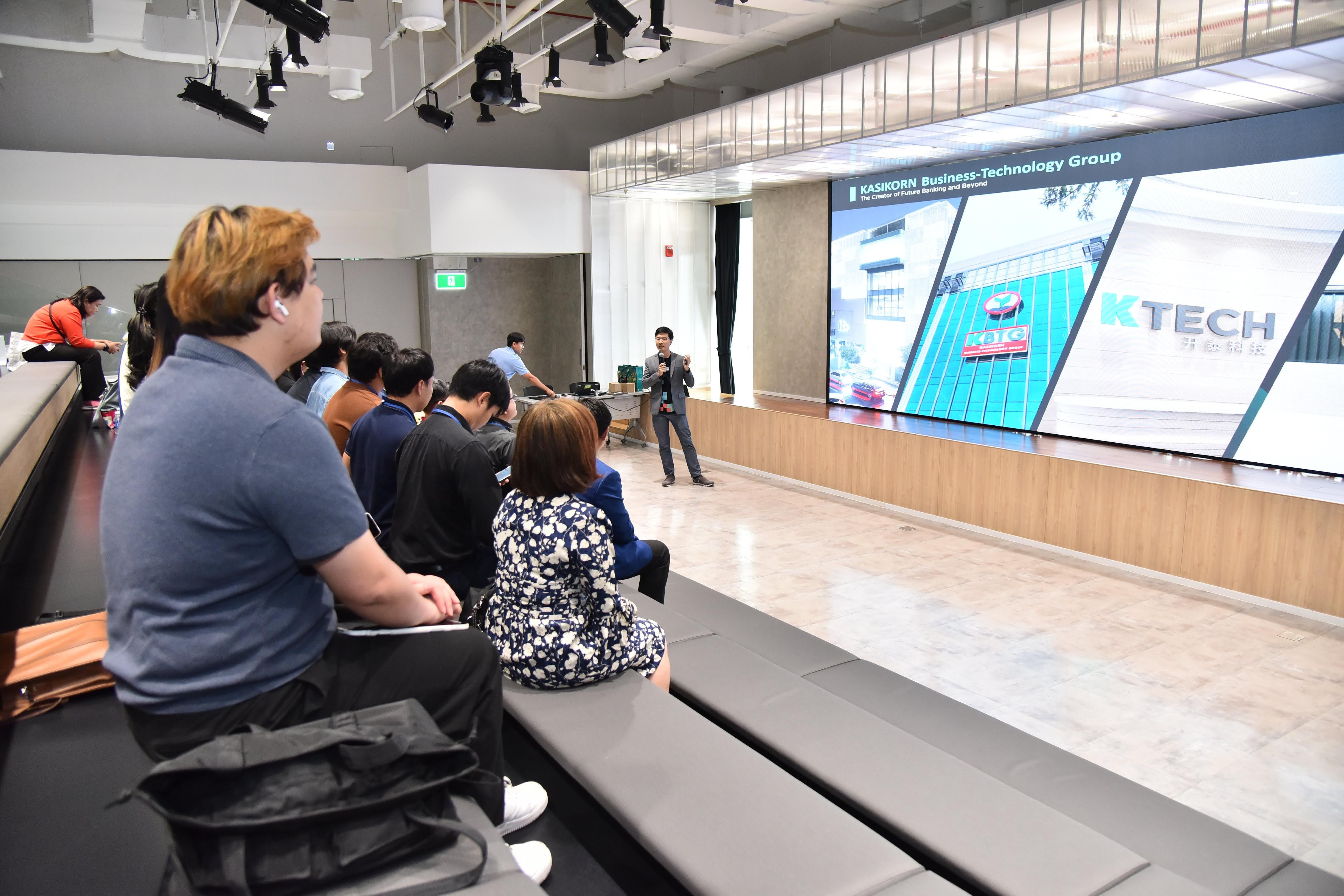 Speaker presents to an audience seated in a hall with a large presentation screen.