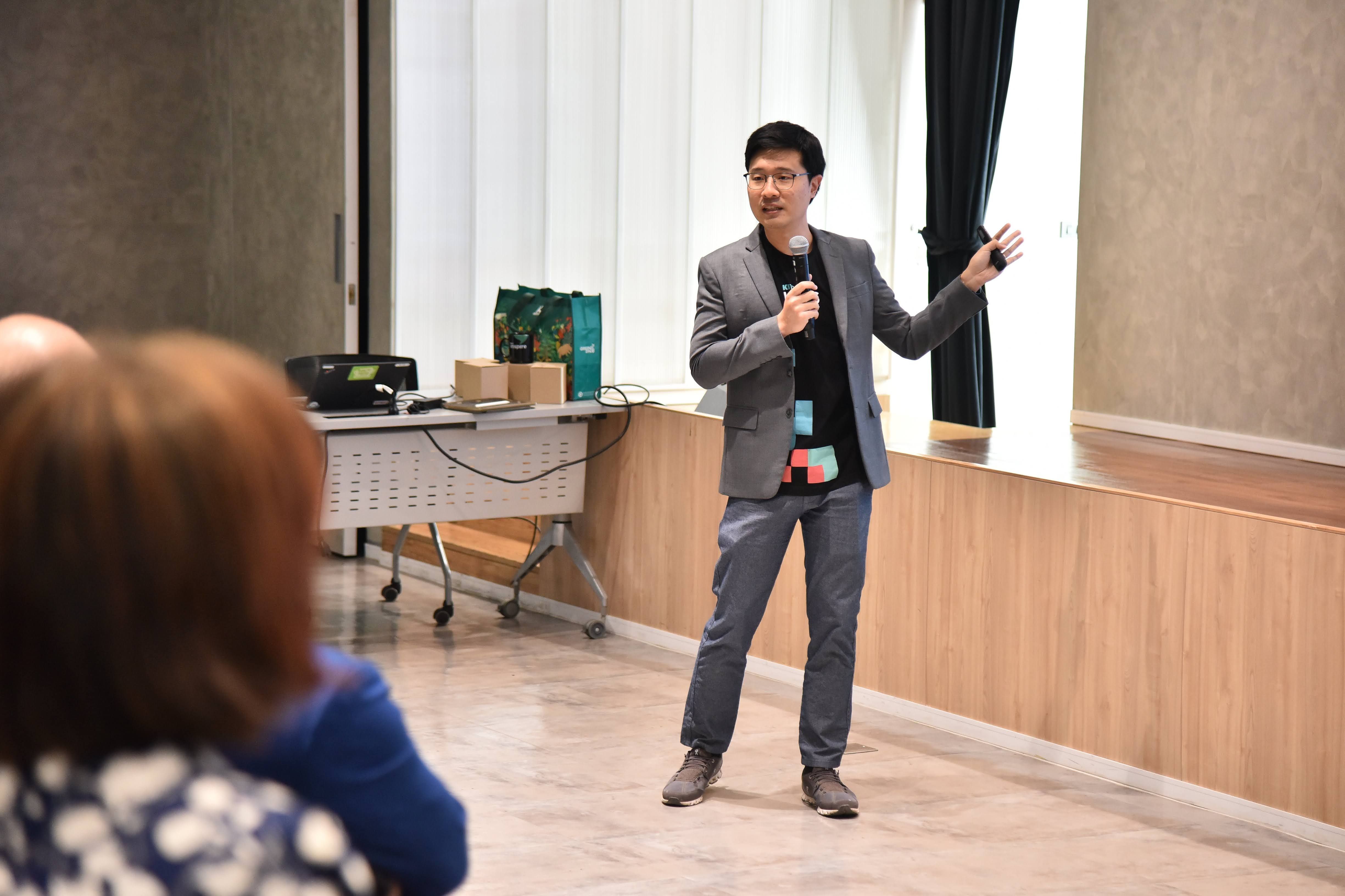 A man in a suit speaks into a microphone, gesturing to an unseen audience.
