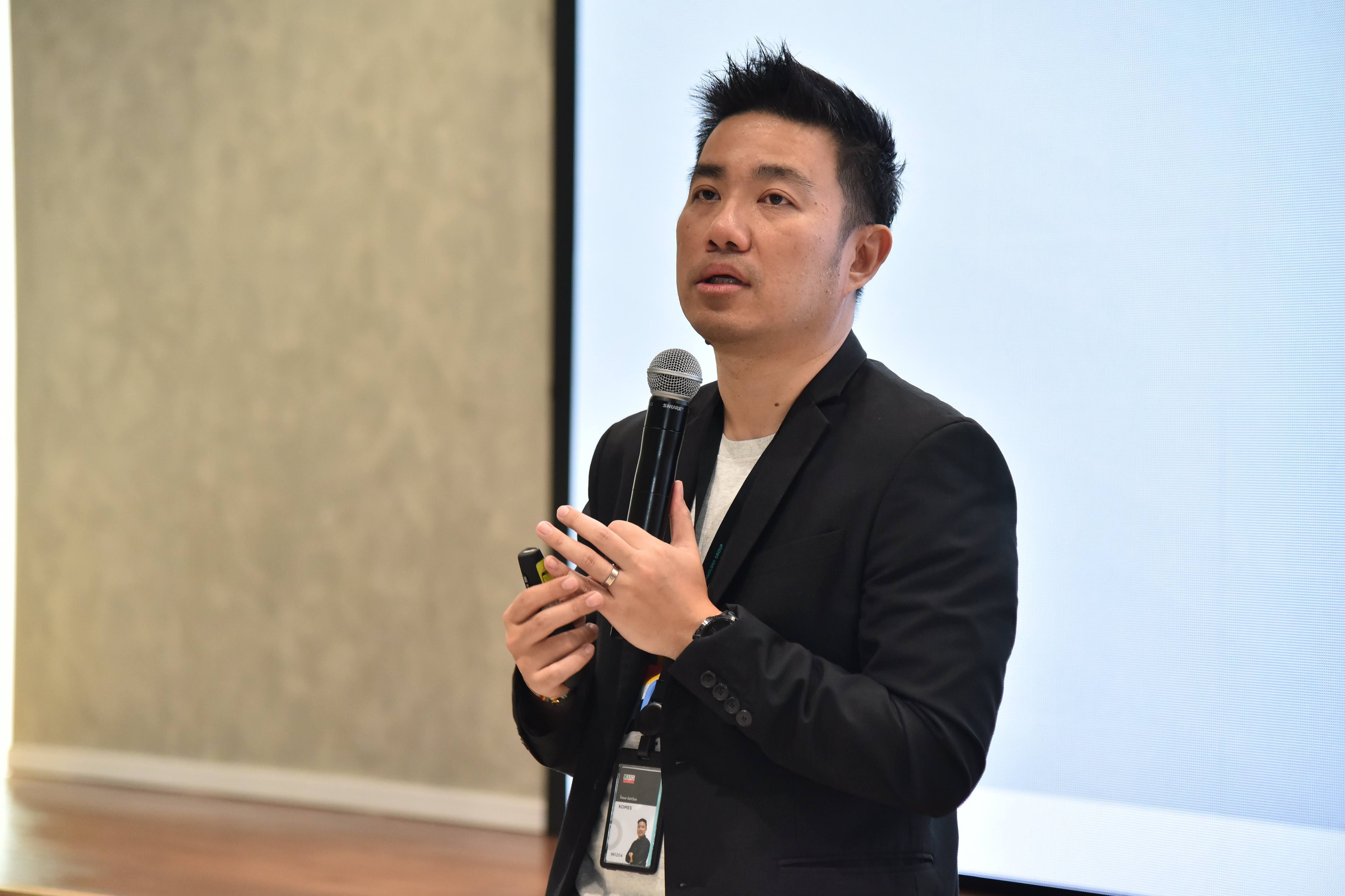 Young Asian man in dark jacket speaks into microphone during presentation.