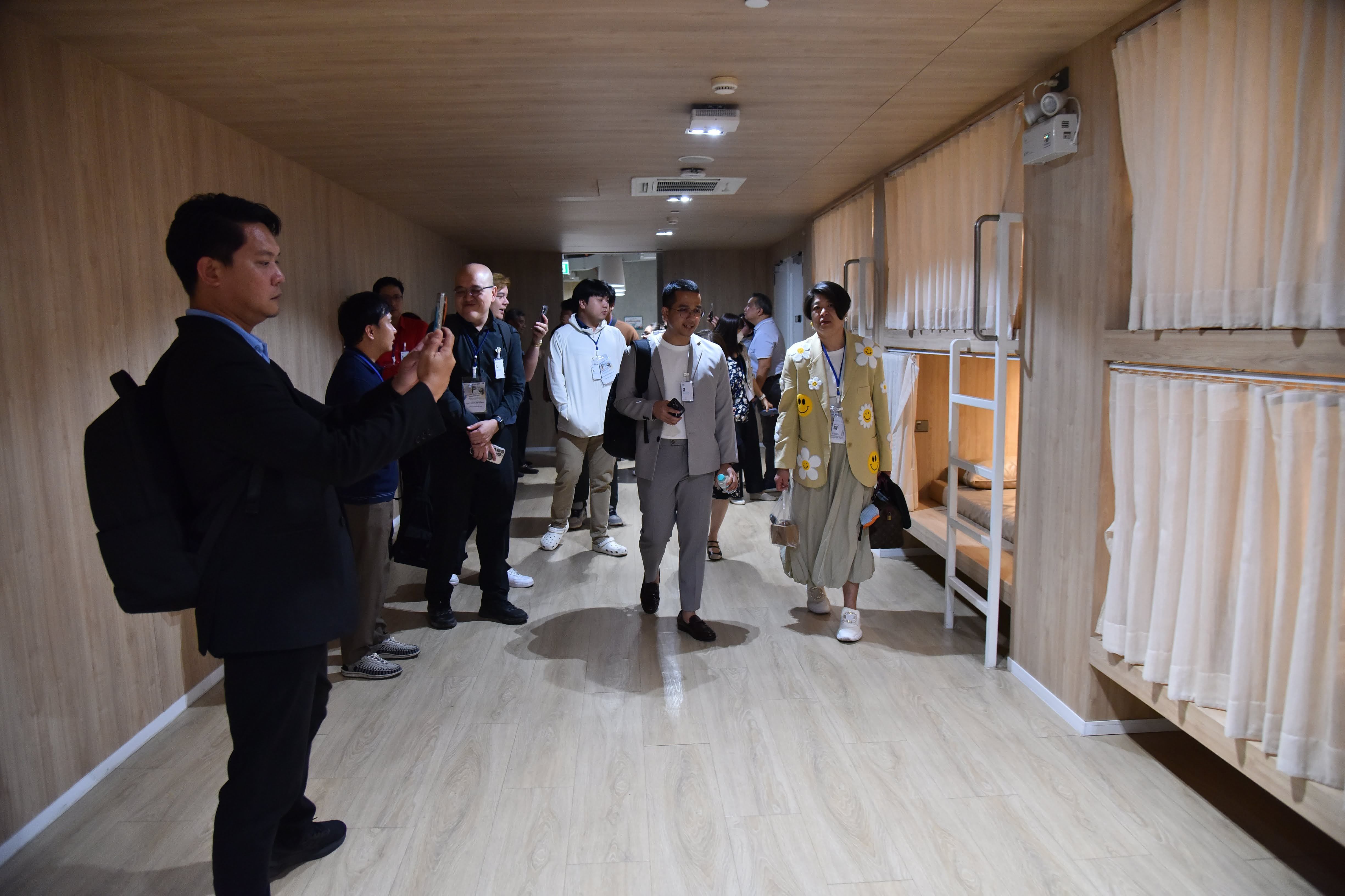 People walk through a narrow, wood-paneled hallway with bunk beds lining one side.