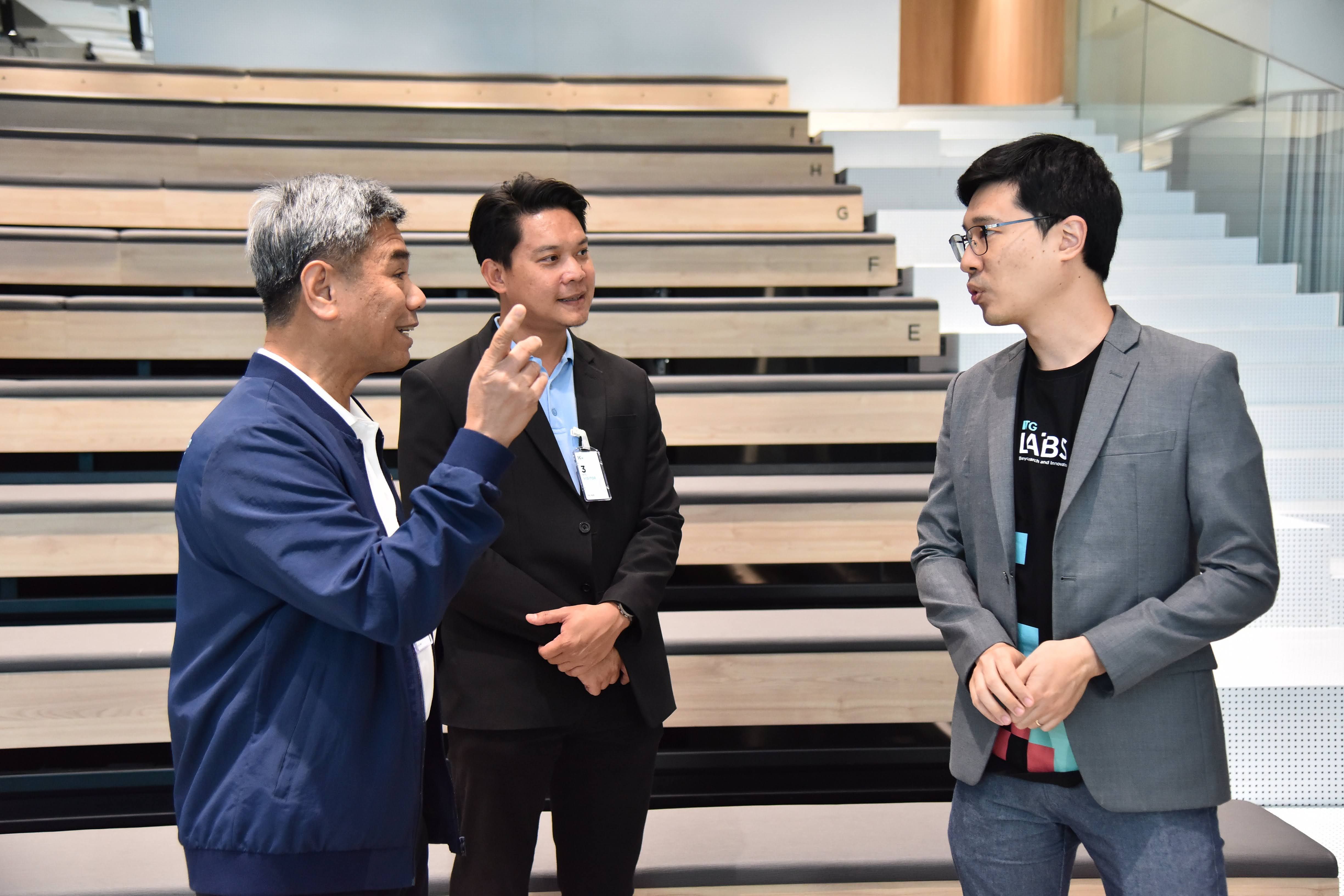 Three men, one gesturing, converse in a modern auditorium setting.