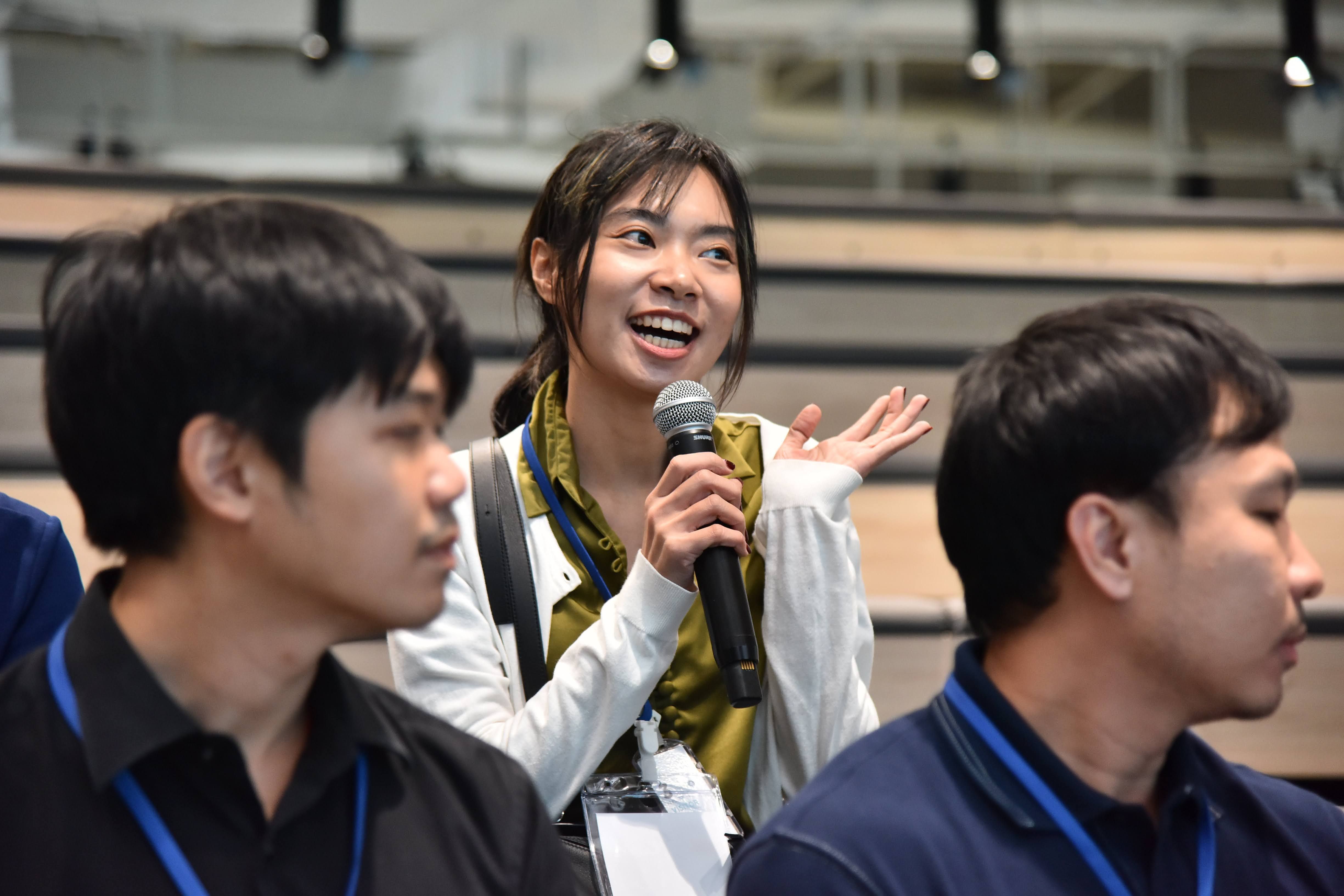Young woman speaking into a microphone, smiling and gesturing in a conference setting.