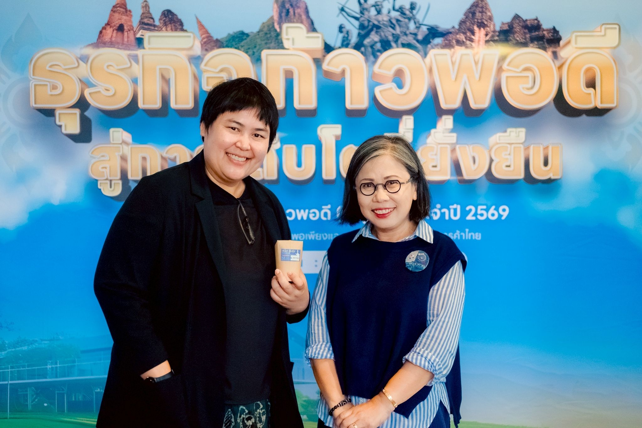 Two smiling women pose in front of a backdrop with Thai text and a sky graphic.