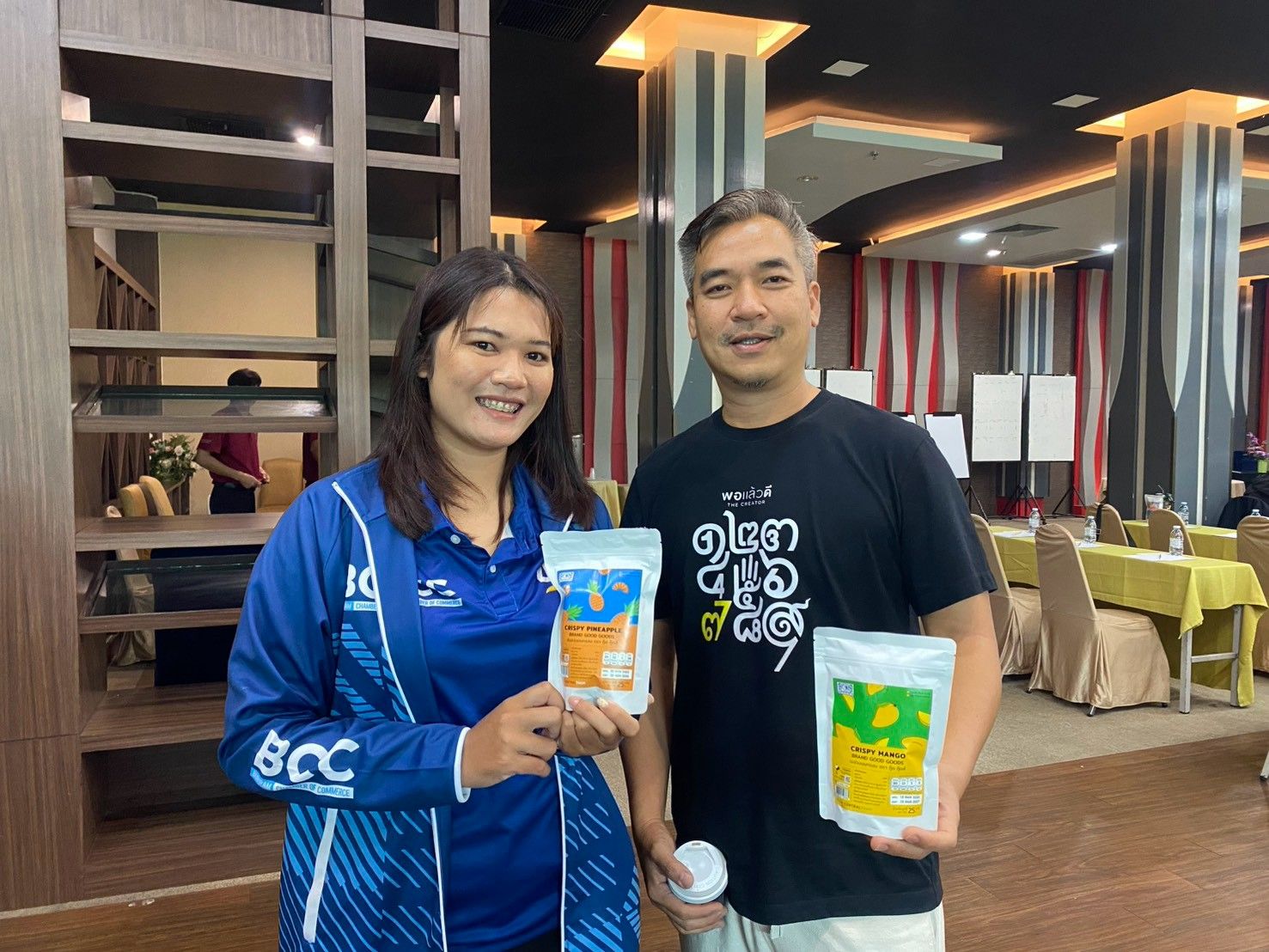 Two people smiling, holding product packages in an indoor event setting.