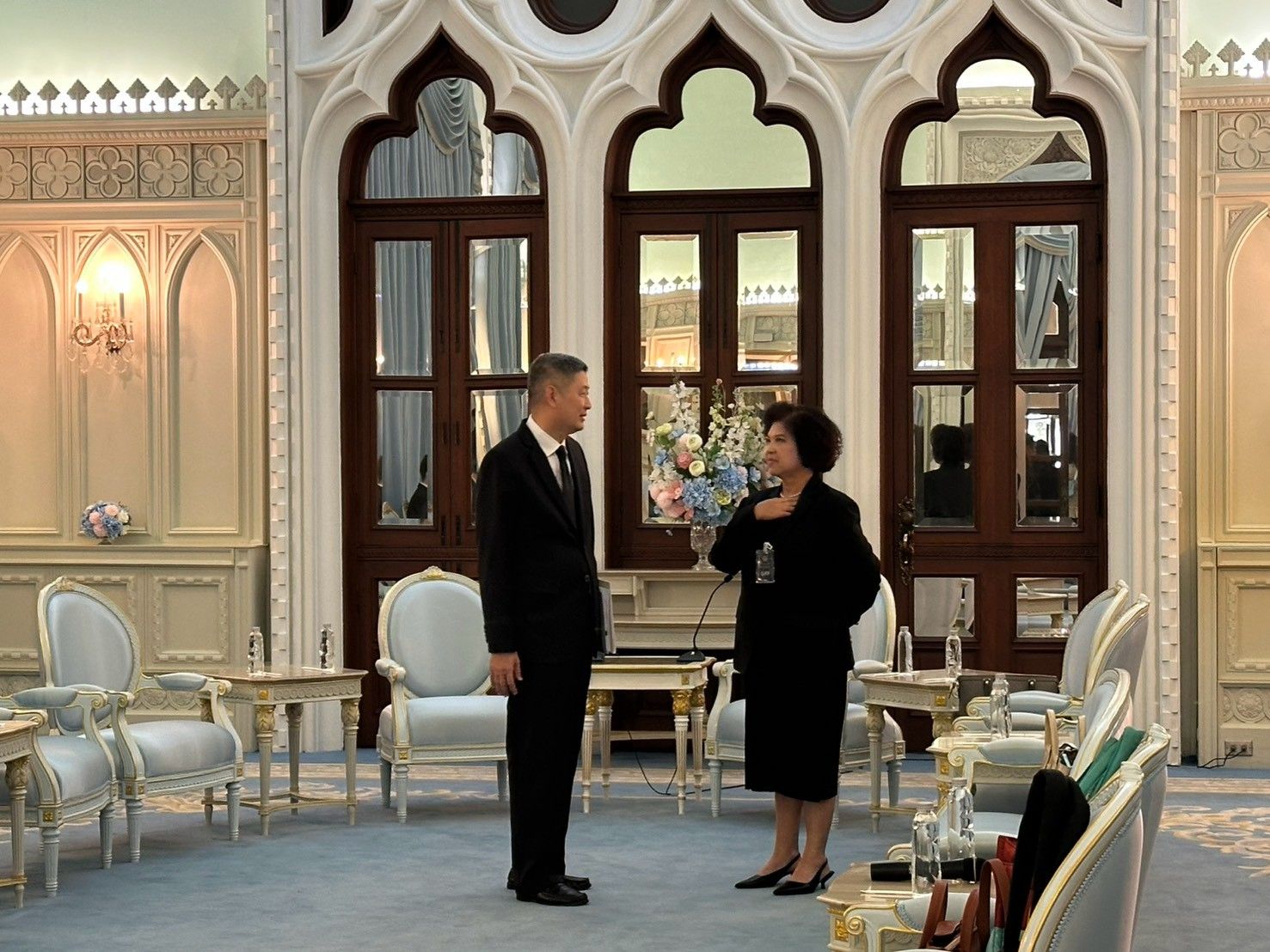 A man and woman in formal attire stand conversing in an ornate, elegant reception room.