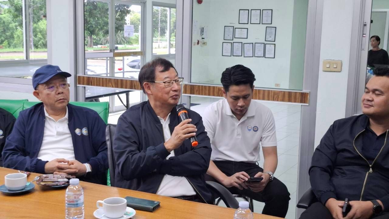 Four men in a meeting, one speaking into a microphone at a table with coffee.
