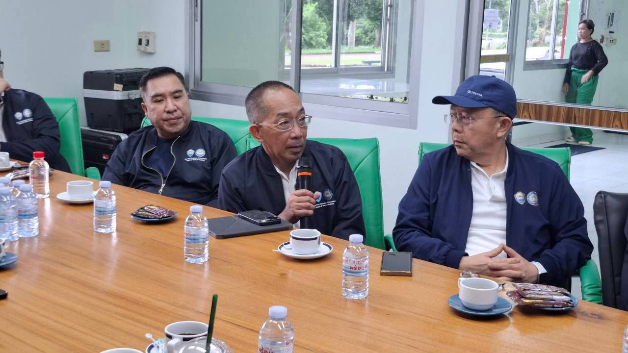 Three men in dark jackets discuss at a conference table, one speaking into a microphone.