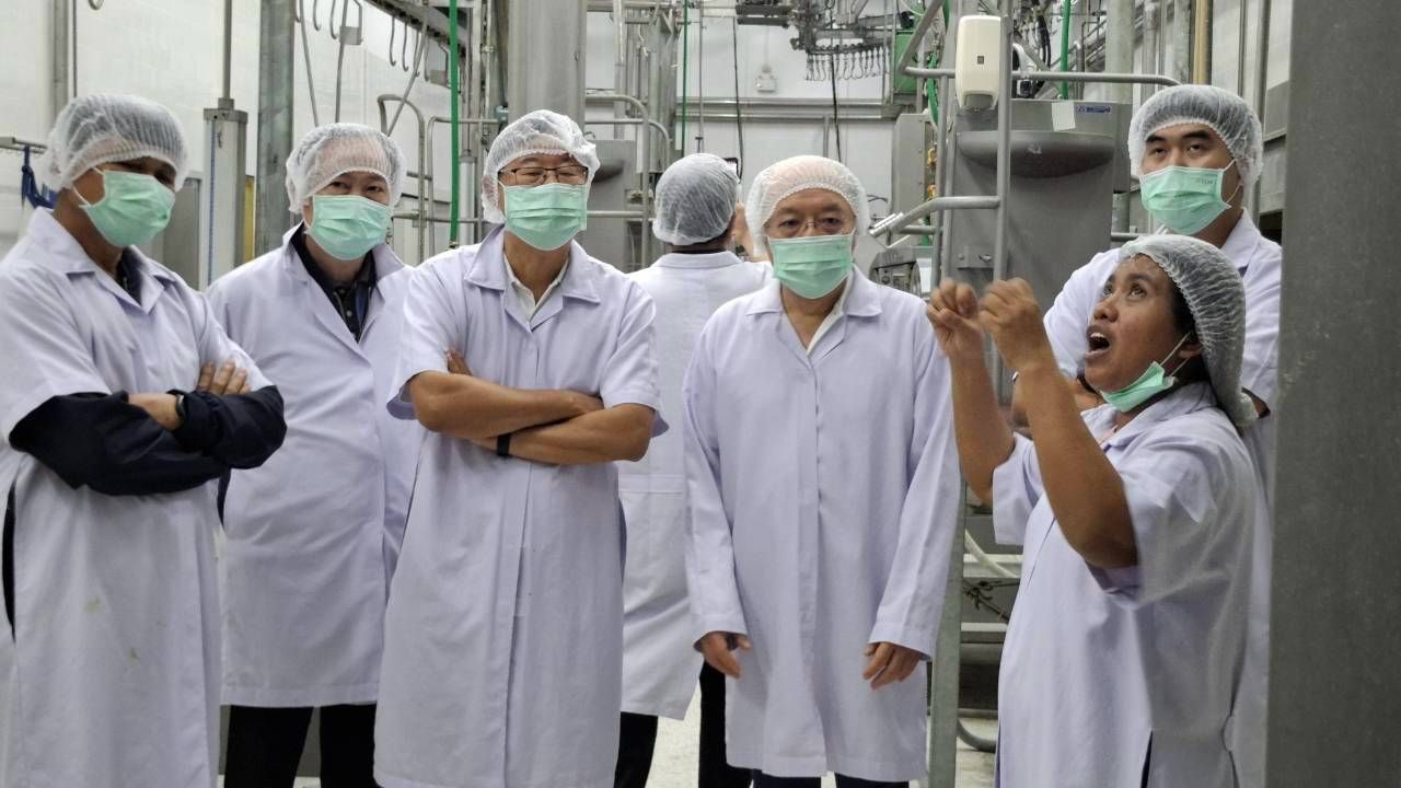Group of masked workers in lab coats listening to a speaker in a sterile factory.